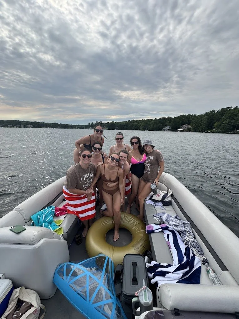 A group of eight women on a boat in a lake, some wearing swimsuits, sunglasses, and hats, with a cloudy sky overhead and trees in the background.