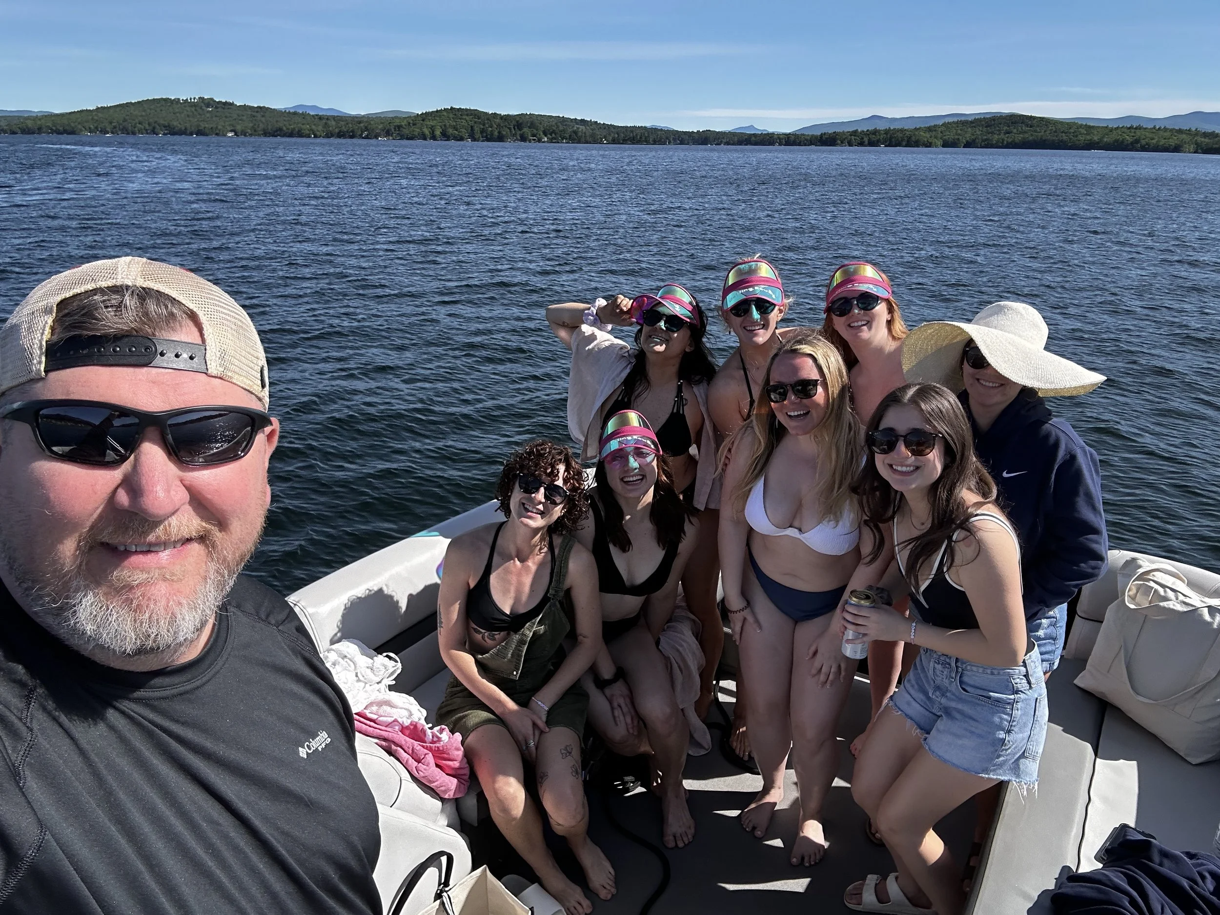 Group of people on a boat taking a selfie with a lake and forested hills in the background.