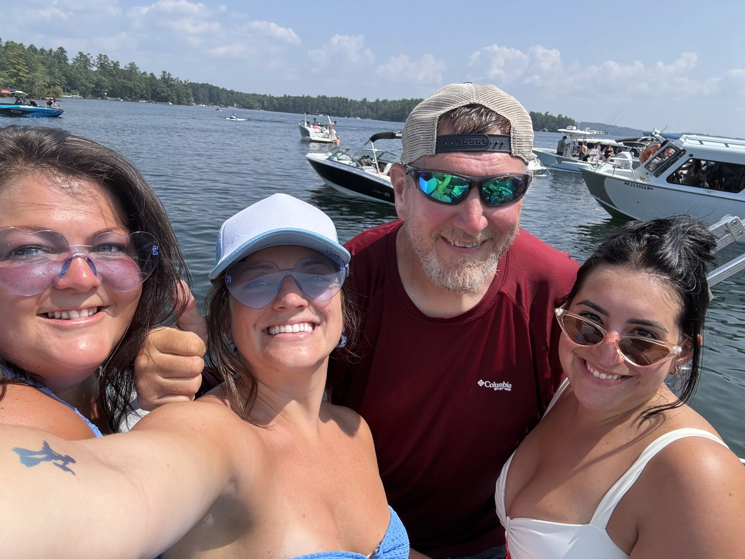 Group of people taking a selfie on a boat, with a lake and multiple boats in the background, during a sunny day with partly cloudy sky.
