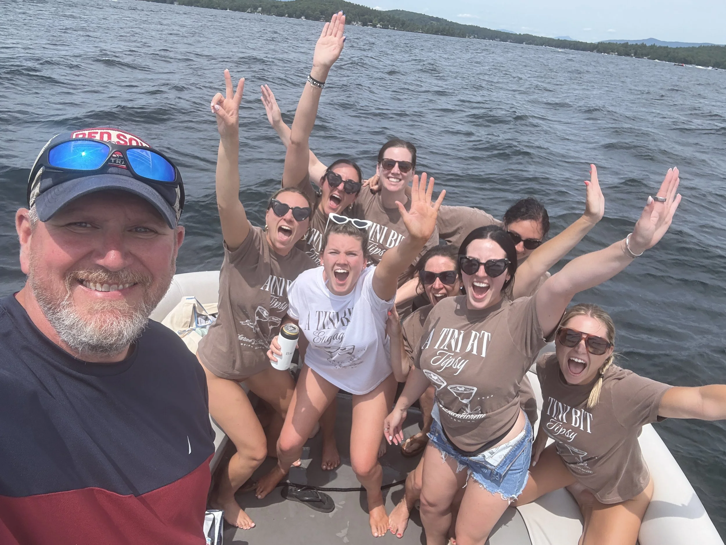 Group of smiling friends on a boat enjoying a lake day, raising their hands and posing for a selfie with water and distant land in the background.