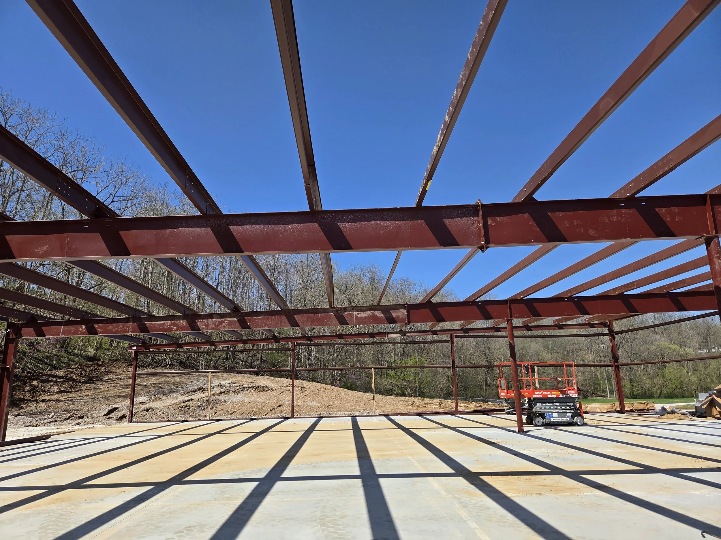Steel framework of a building under construction on a sunny day with clear blue sky, with trees in the background and shadow lines cast on the ground.