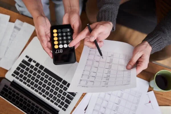 People analyzing data charts on paper, one person holding a calculator and a pen, with laptops and a coffee mug on the desk.