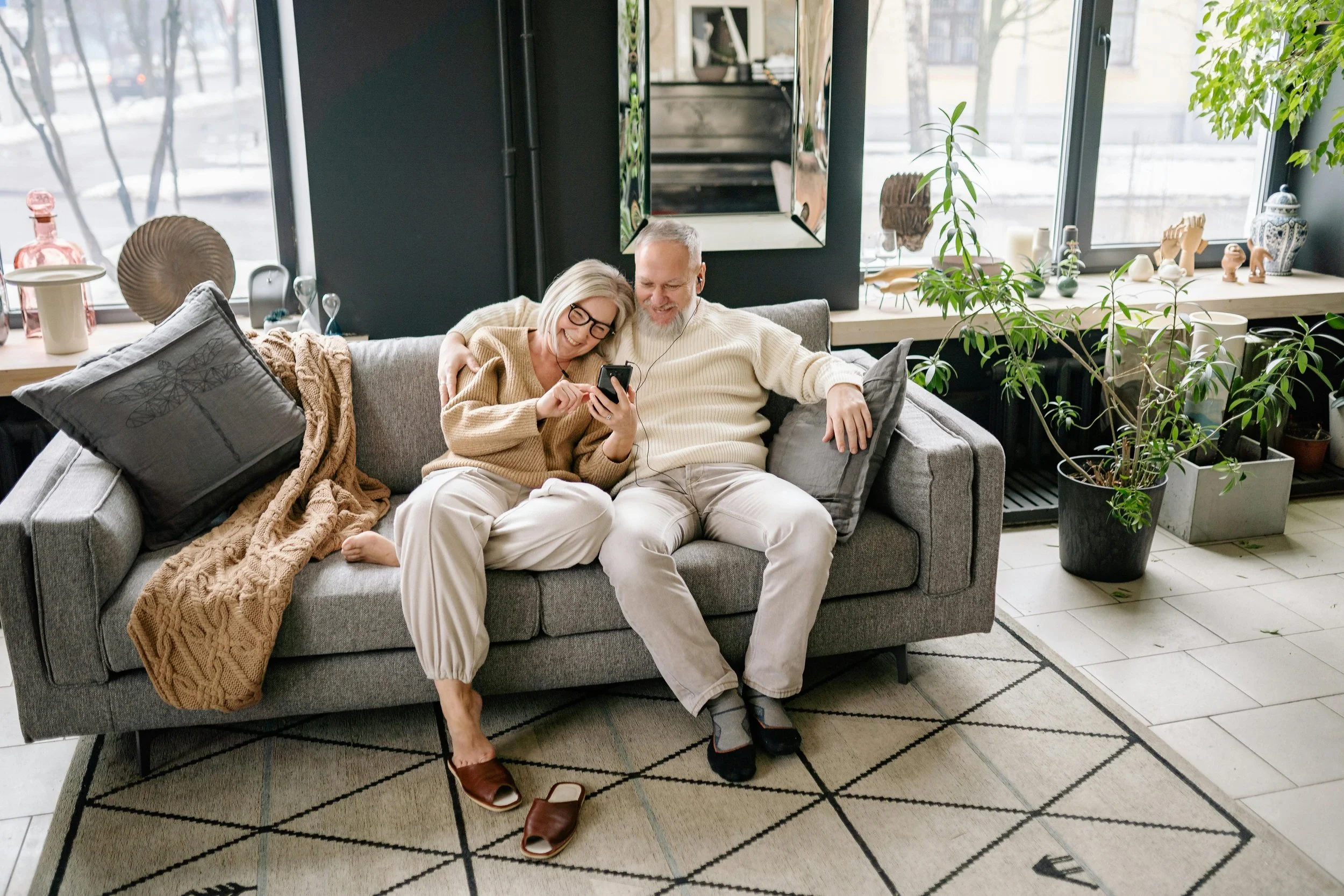 A smiling elderly couple sitting on a gray sofa, looking at a smartphone together in a modern living room with large windows, houseplants, and decorative items on the windowsill.