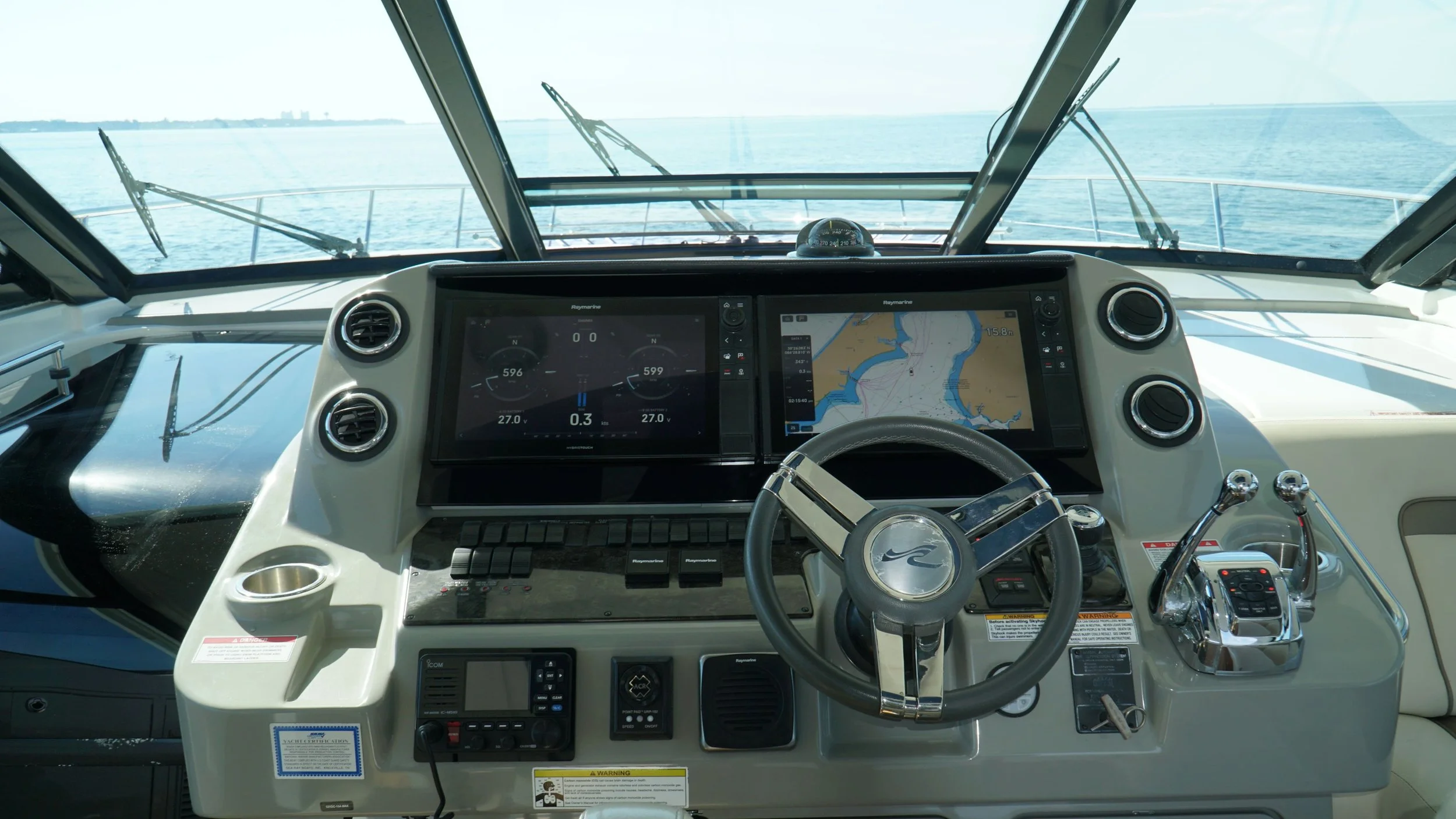 The image shows the interior view of a boat's helm station with navigation and control equipment, looking out over the water with a distant shoreline.