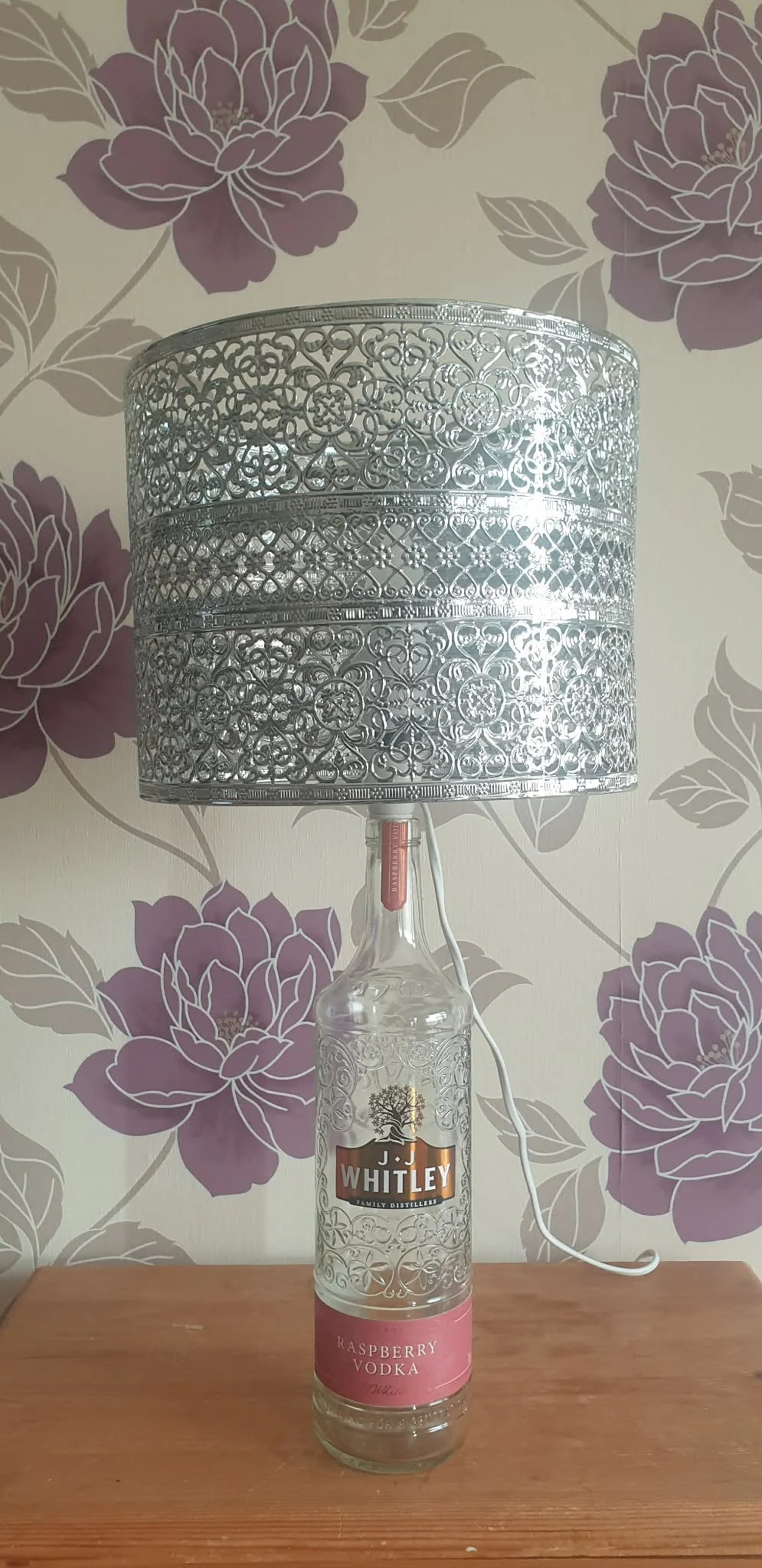 A lamp made from a clear raspberry vodka bottle with a decorative silver lampshade on top, placed on a wooden surface against a floral wallpaper background.
