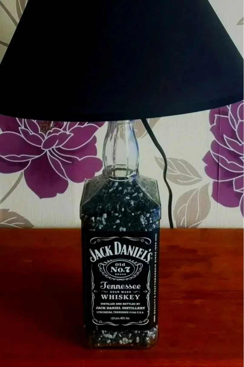 A table lamp made from a Jack Daniel's Tennessee whiskey bottle with a black lampshade, positioned in front of floral wallpaper.