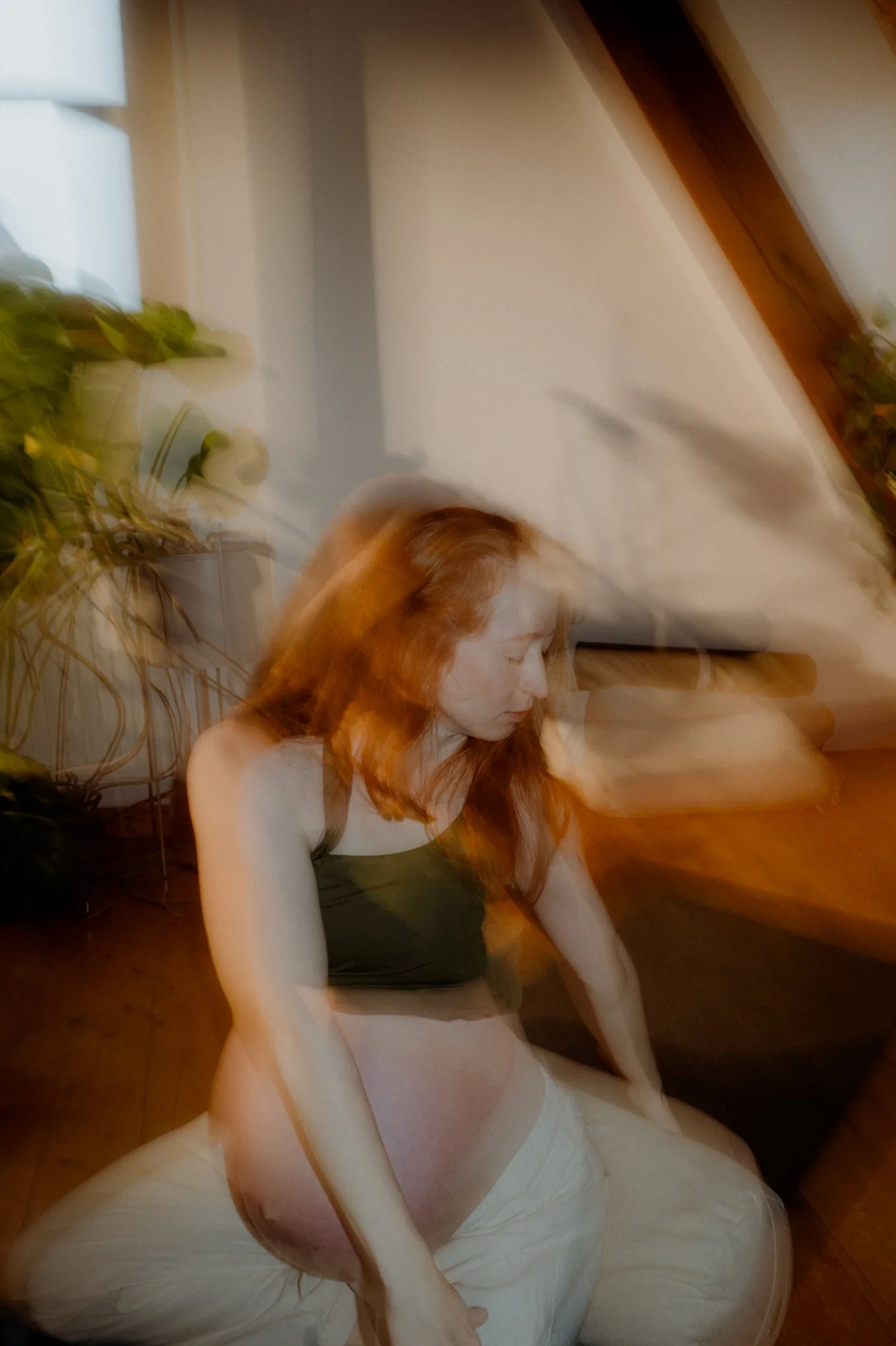 A woman with long red hair wearing a gray tank top and white skirt, sitting on the floor with her eyes closed, in a room with wooden beams, a white wall, and a potted plant.
