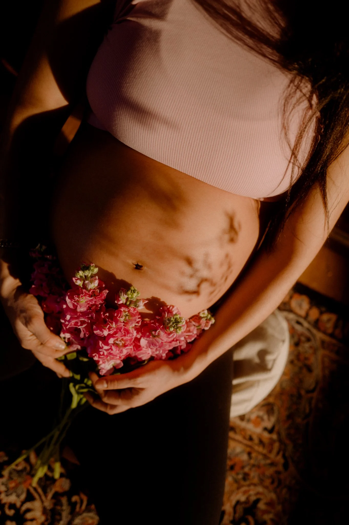 A woman holding a bouquet of pink flowers, wearing a pink top and black pants, with a belly button piercing, sitting on a patterned rug.