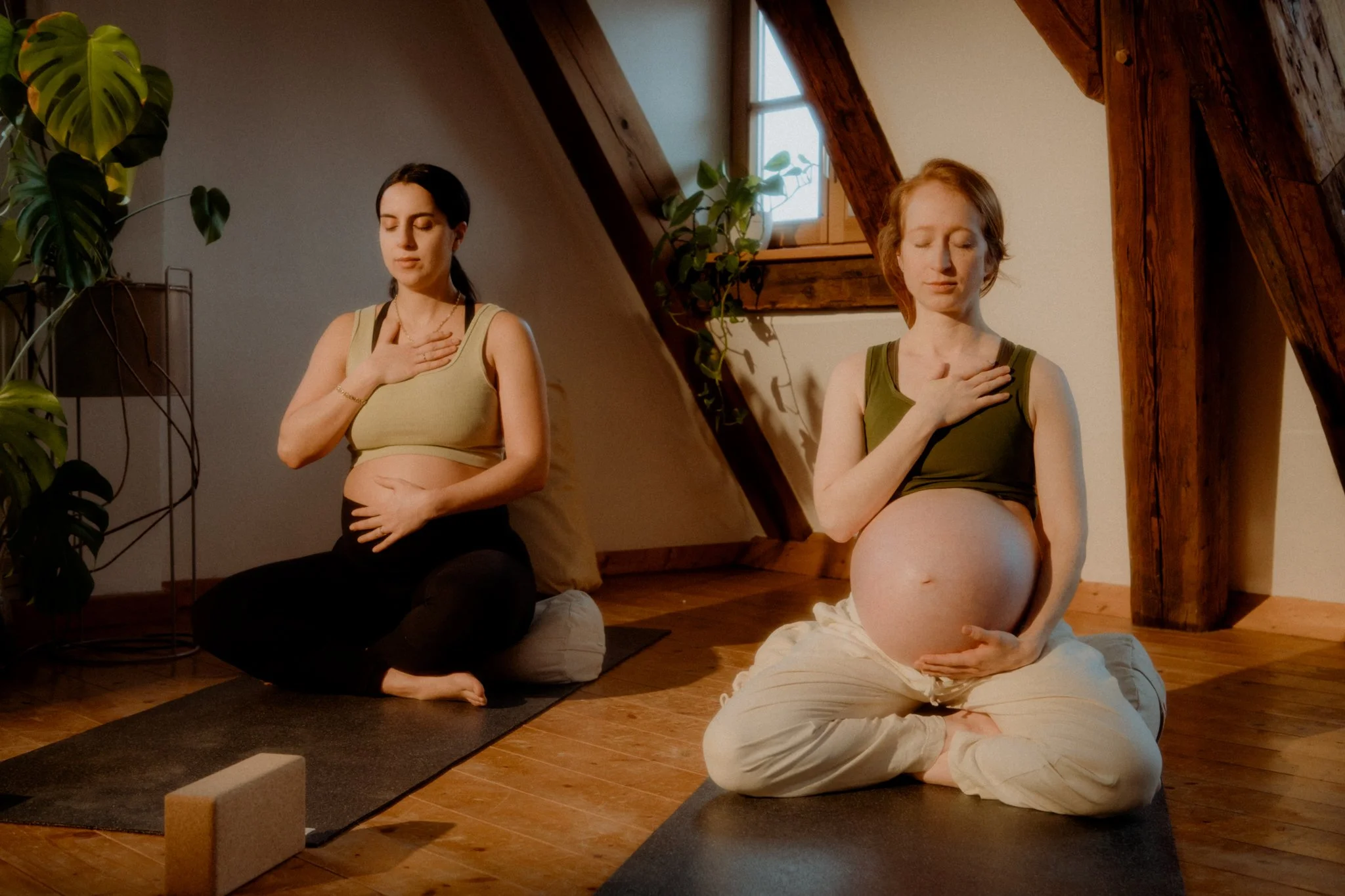 Two women practicing yoga indoors; one pregnant woman and one with her hand over her chest, eyes closed, seated on a yoga mat in a room with wooden beams and a slanted ceiling, natural light coming through a window, surrounded by plants.