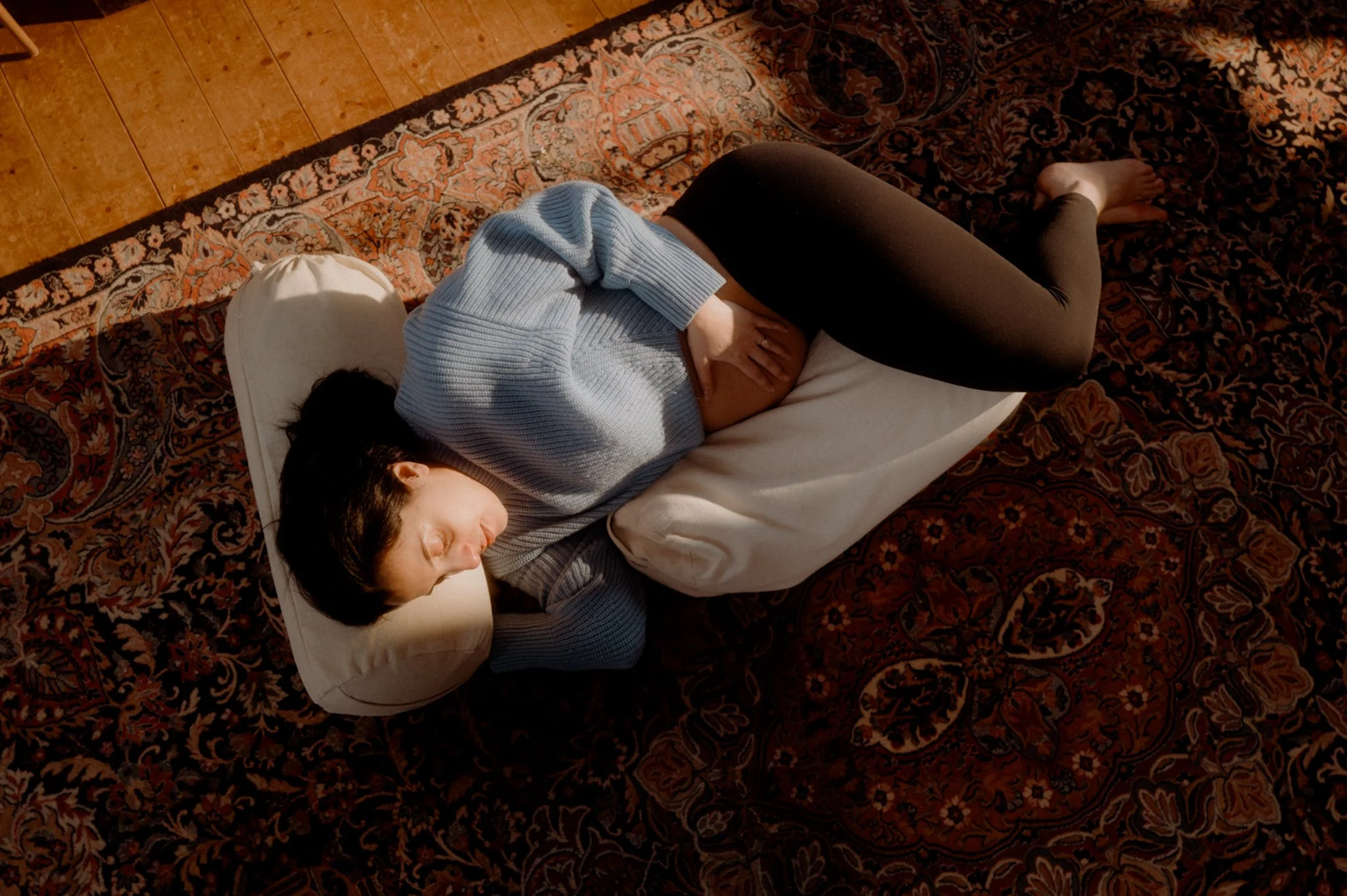 A woman lying on her side on a bed with patterned bedding, wearing a white sweater and black leggings, with her head resting on a pillow.