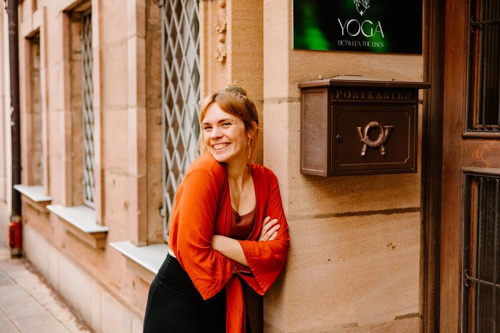 A woman smiling and leaning against a stone wall outside buildings with barred windows, wearing a red jacket and black skirt.