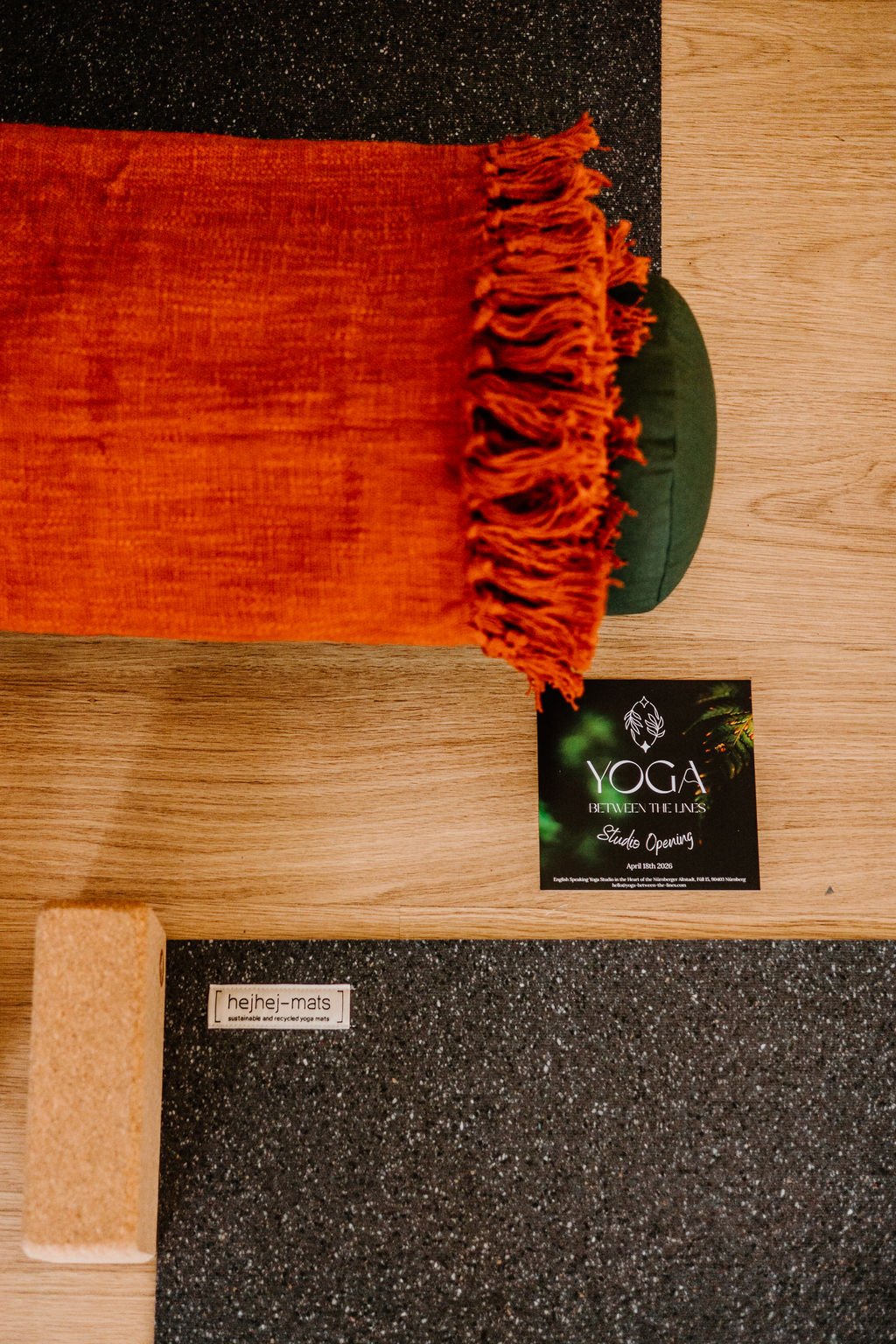 A yoga studio display with an orange fringed yoga mat, a green cushion, a cork yoga block, and a black card with green and white text about yoga studio opening.