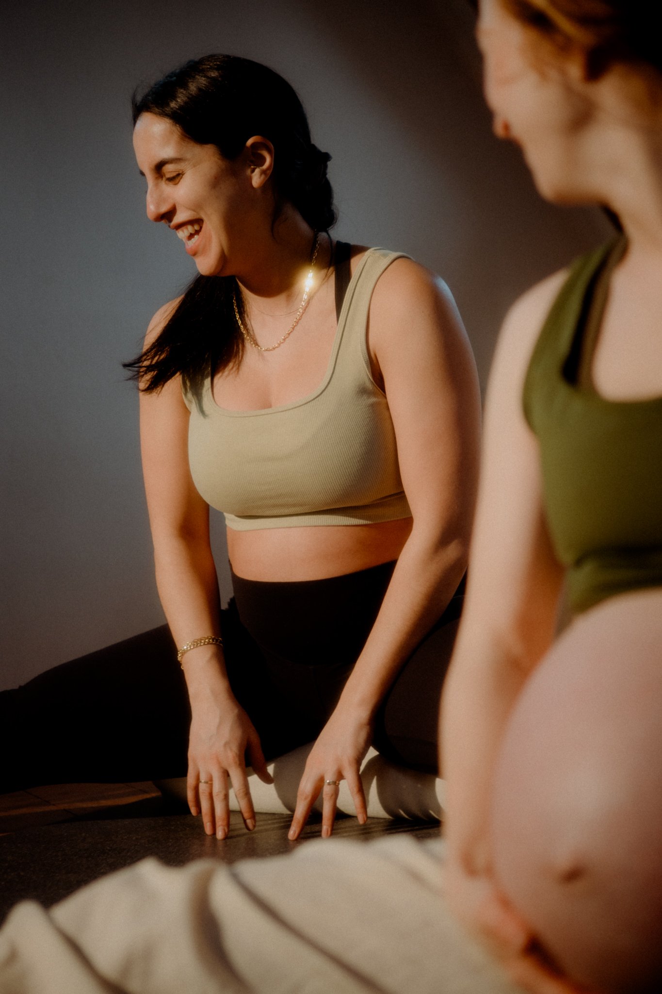 Two women practicing yoga indoors, one smiling with eyes closed, wearing a beige sports bra and black leggings, the other partially visible with a similar outfit, both in a forward bend position with hands on the floor.