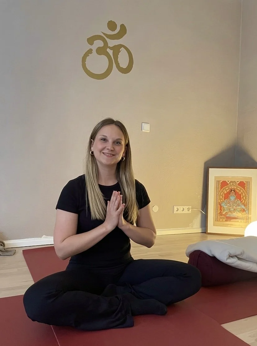 A woman practicing yoga in a peaceful room, sitting cross-legged on a yoga mat with hands in a prayer position, smiling at the camera.