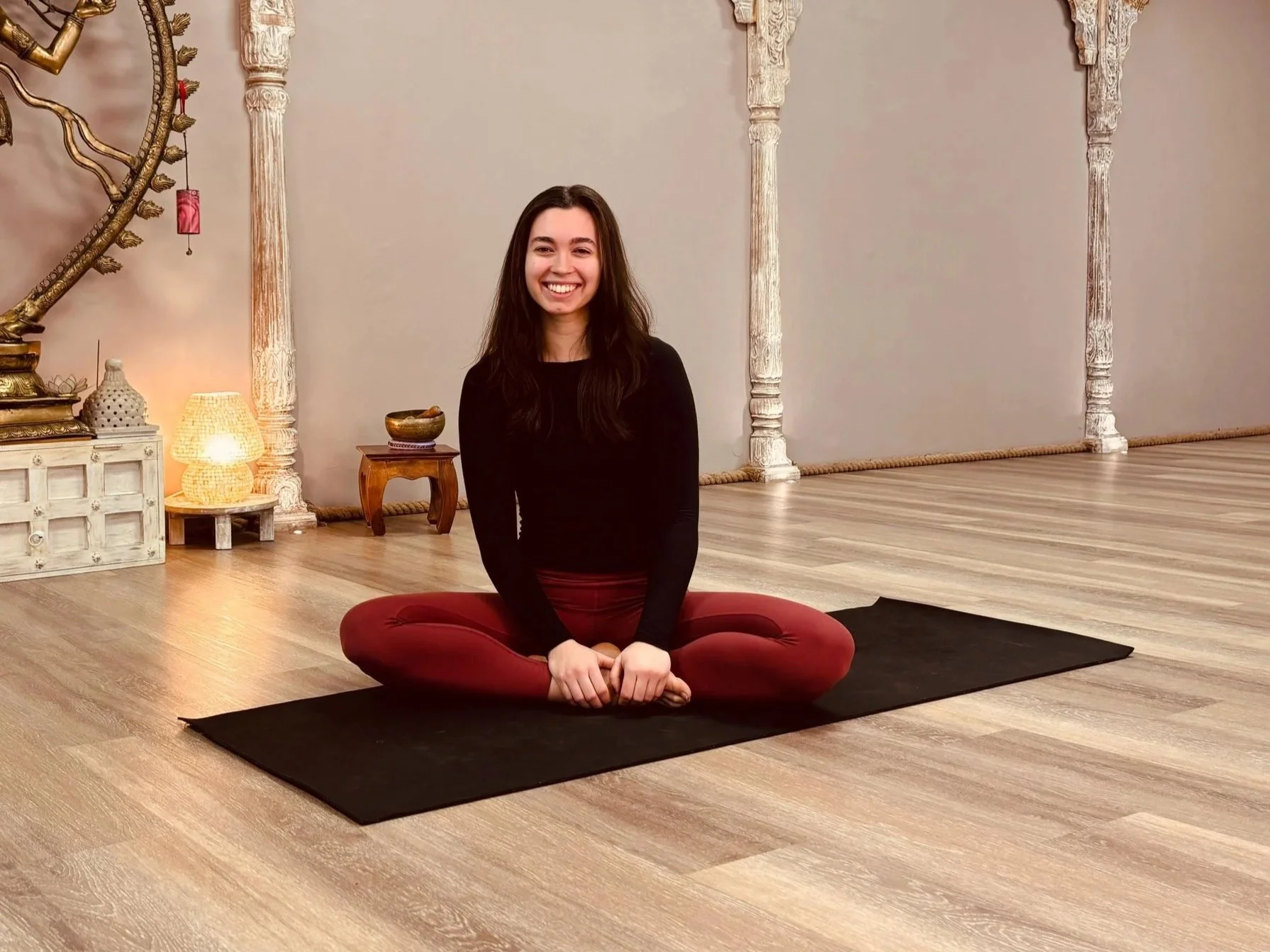 A young woman sitting on a yoga mat with her legs crossed in a yoga studio, smiling at the camera, wearing a black top and burgundy yoga pants, with a serene background of decorative pillars, lamps, and a wooden floor.