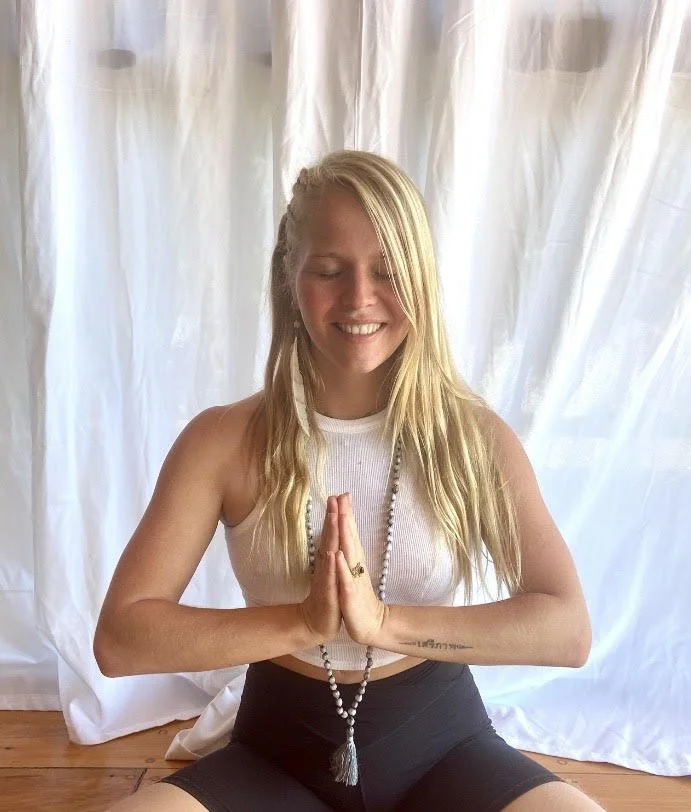 Young woman with long blonde hair sitting cross-legged in front of white curtains, smiling with eyes closed, hands in prayer position.