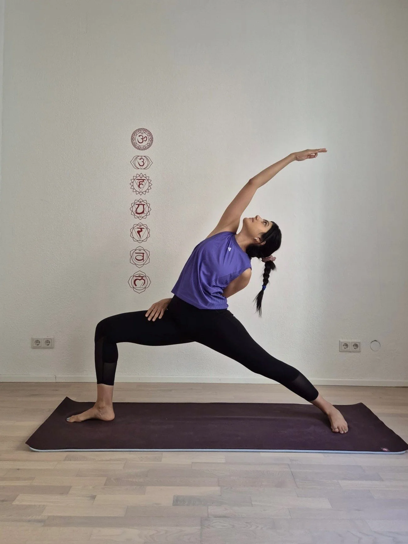 A woman wearing a purple tank top and black leggings practicing yoga in a lunge pose on a mat in a studio. The wall behind her has chakra symbols painted vertically.