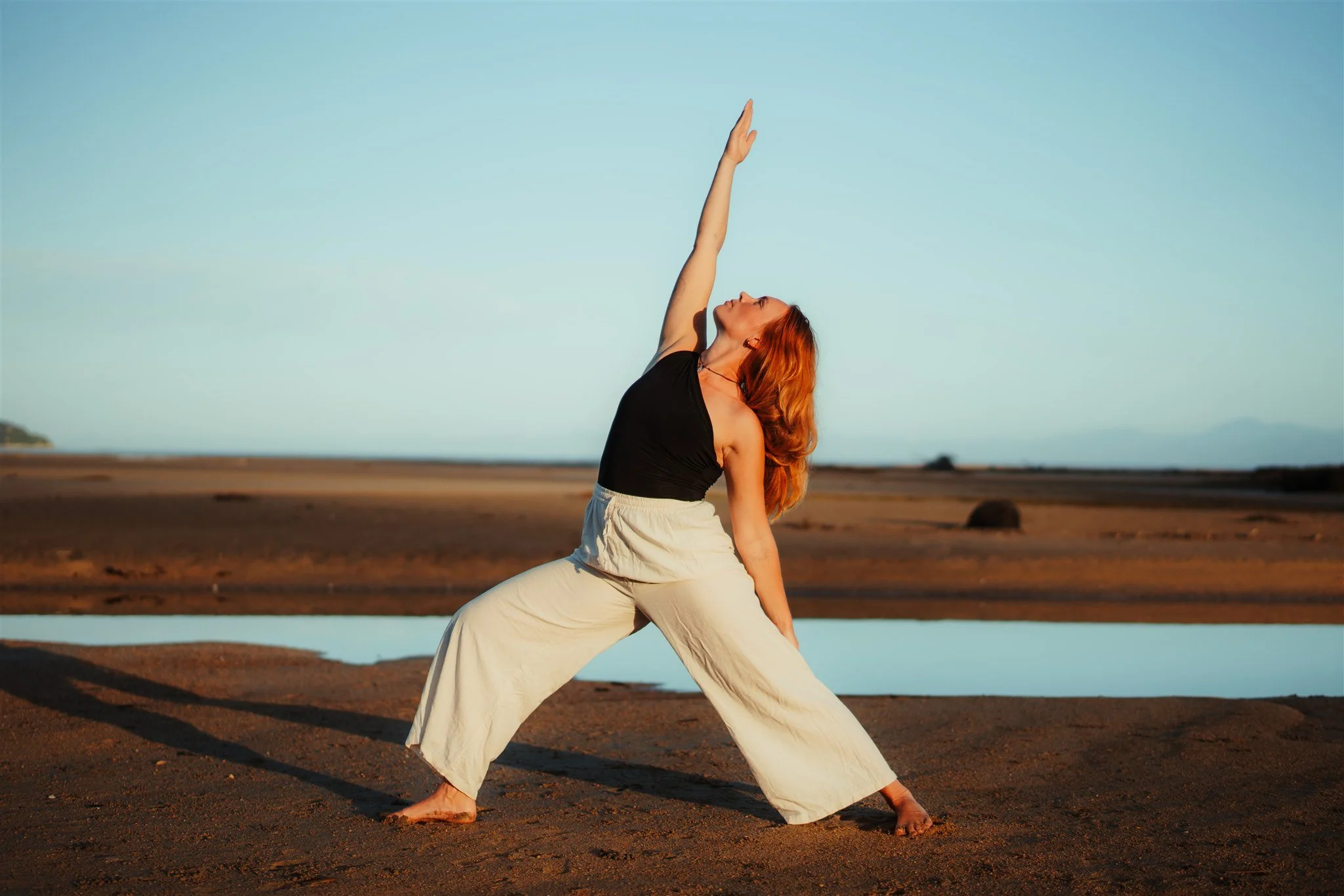 A woman practicing yoga on a beach during sunset, stretching with one arm raised towards the sky.