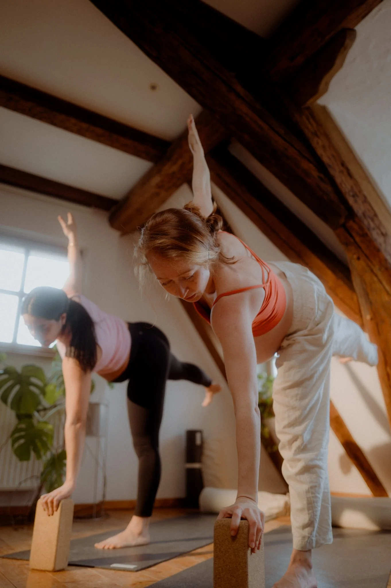 Two women practicing yoga in a studio, balancing on yoga blocks with one arm extended upward and the other hand on the blocks, with wooden beams on the ceiling and a large window.