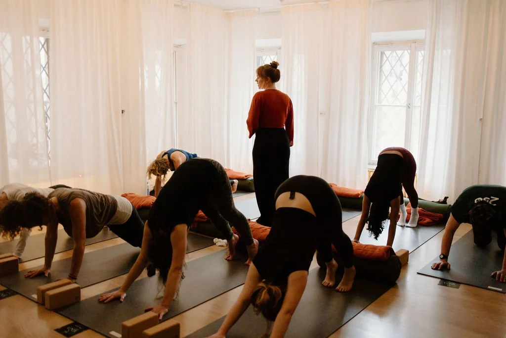 A group yoga class in a bright, airy room with white curtains, with a yoga instructor standing at the front guiding students through a pose.