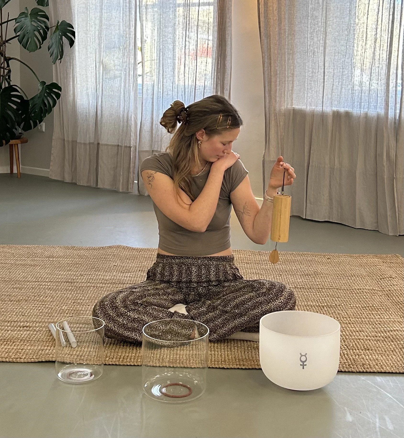 A woman with long hair tied back, wearing glasses, earrings, a gray top, and patterned pants, is sitting on a woven rug in a room with sheer curtains. She is holding a wooden percussion instrument with a string and a small bell. In front of her on a table are three bowls, a white singing bowl with a symbol, and two smaller bowls, one containing a stick and the other possibly a bracelet. There are glass cups on the table, and a large green plant in the background.
