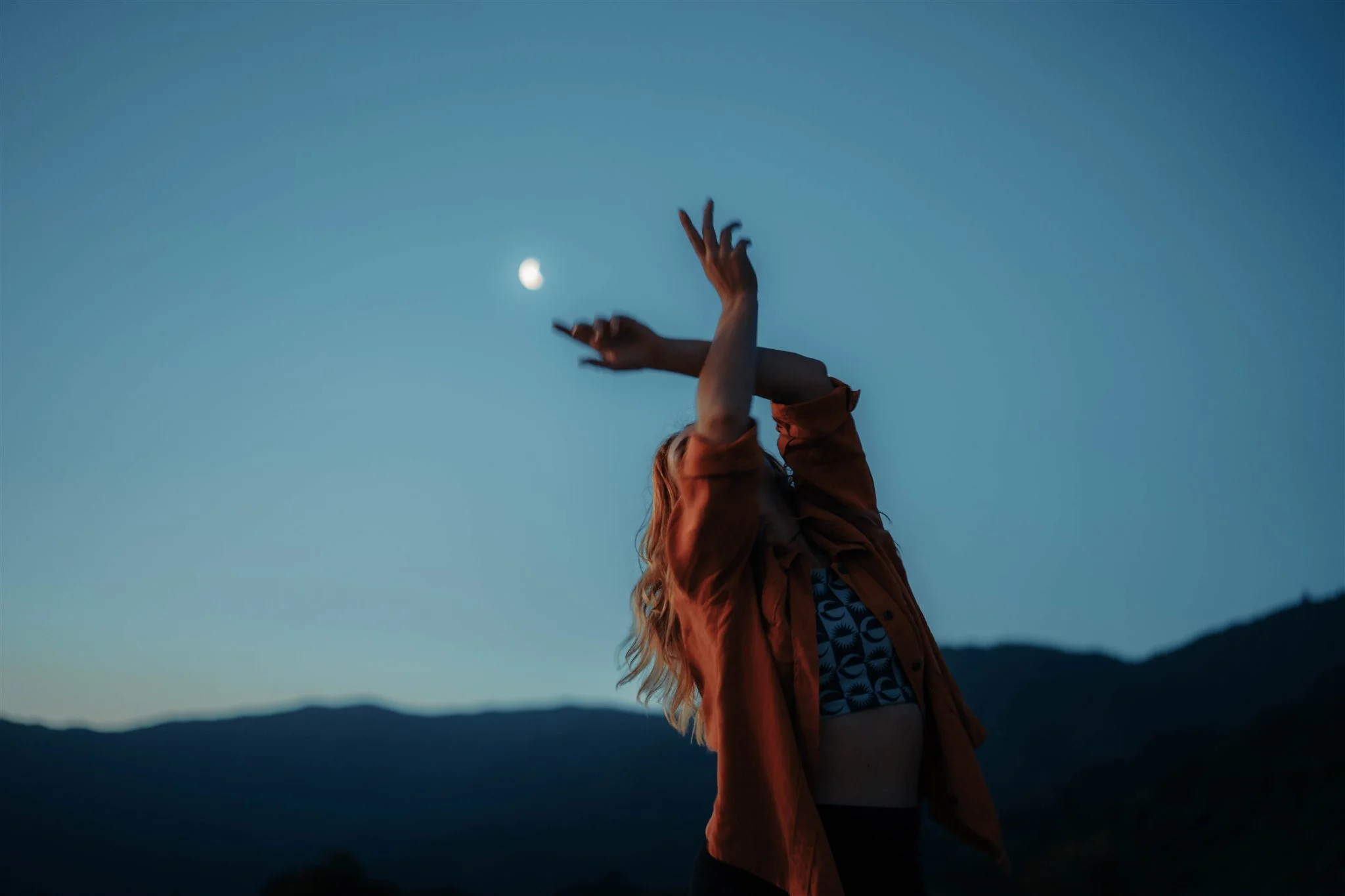 Person with long hair wearing an orange jacket standing outdoors at dusk, reaching toward the sky where the moon is visible.