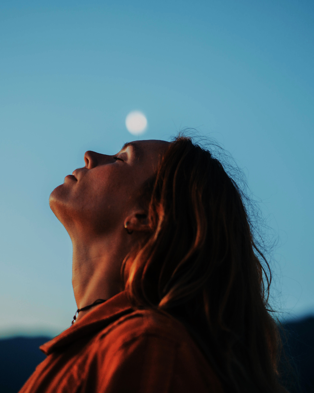 A woman with long hair is outdoors at dusk or dawn, her head tilted back with eyes closed, as the moon rises in the sky behind her.