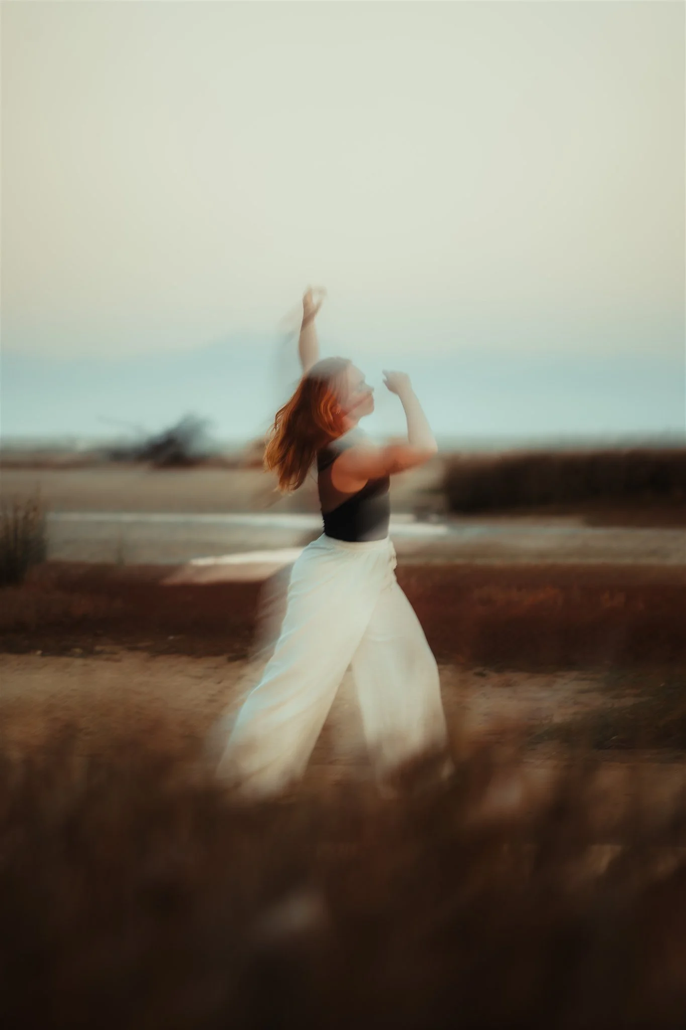 A woman in a black top and white skirt dancing on a beach during sunset, with a blurred background of ocean and sky.