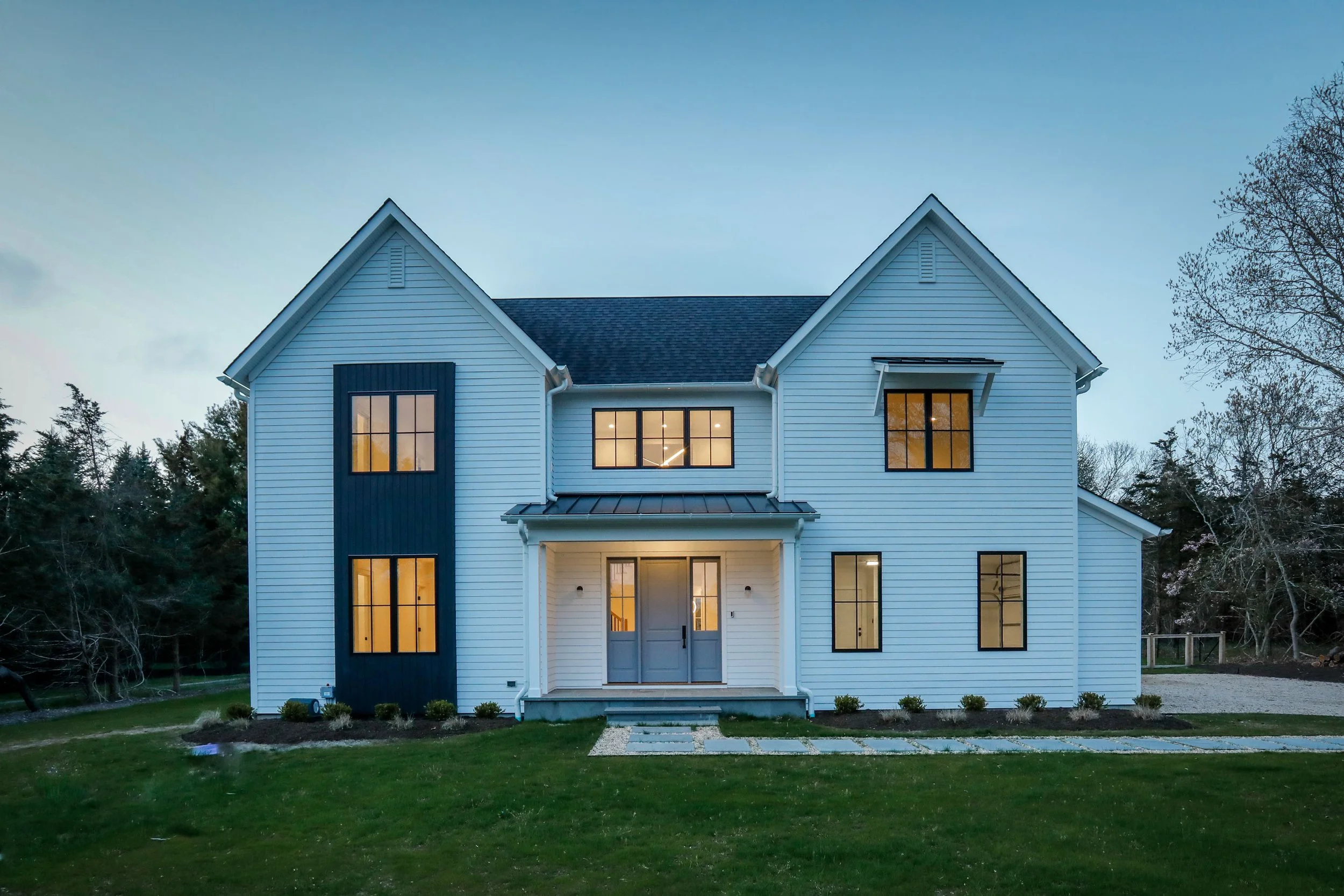 A modern two-story white house with black window frames, front steps, and a front lawn during dusk.