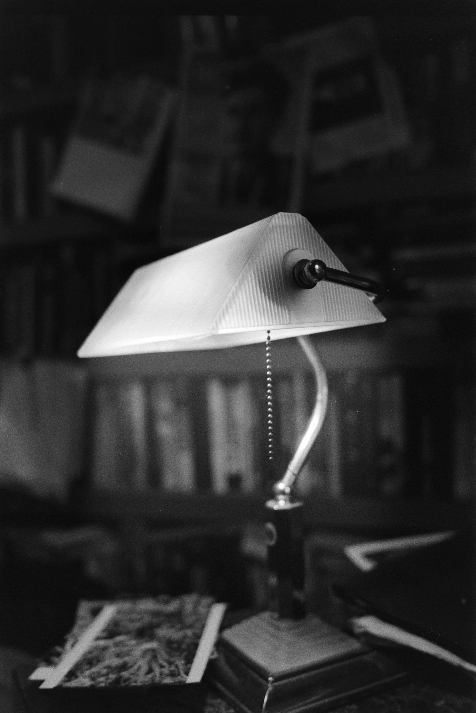 A desk lamp with a bendable arm and a pull chain switch, placed on a table with papers and books in a room with shelves and chairs in the background.