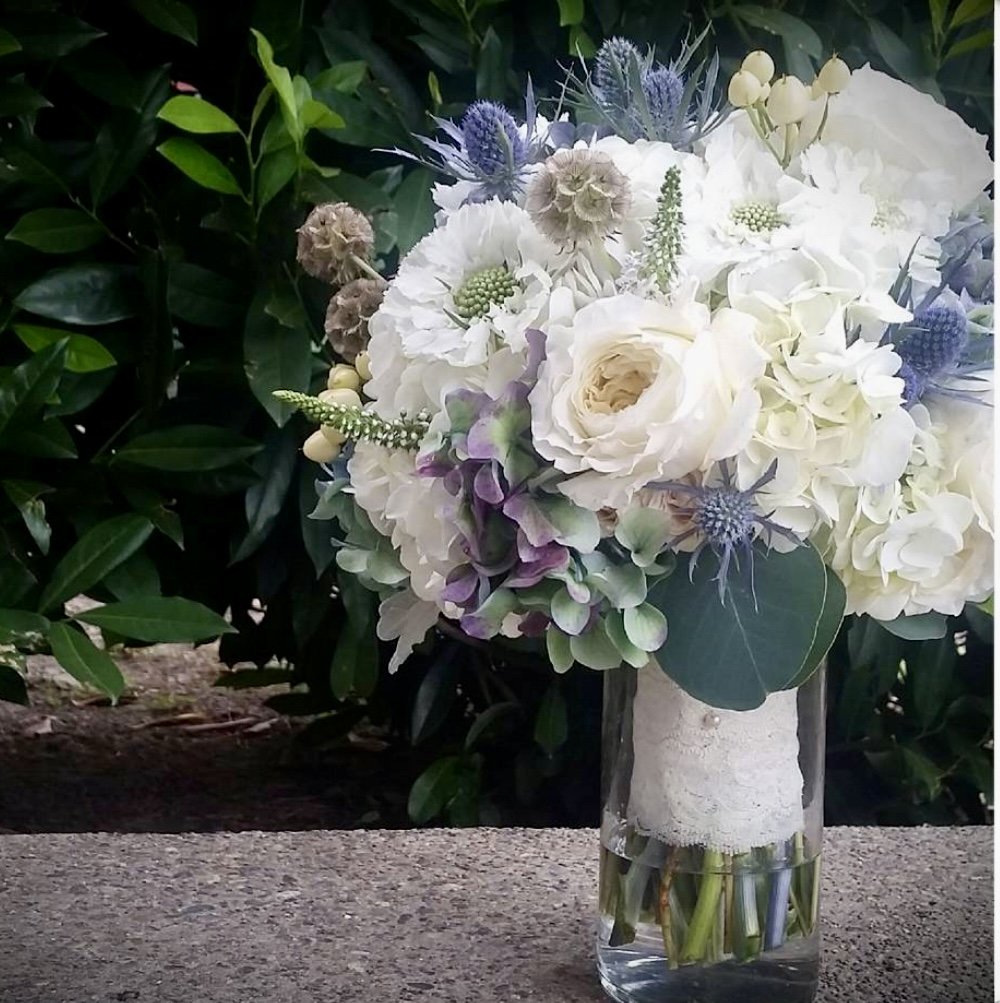 A bouquet of white, purple, and light green flowers in a glass vase on a table outdoors.