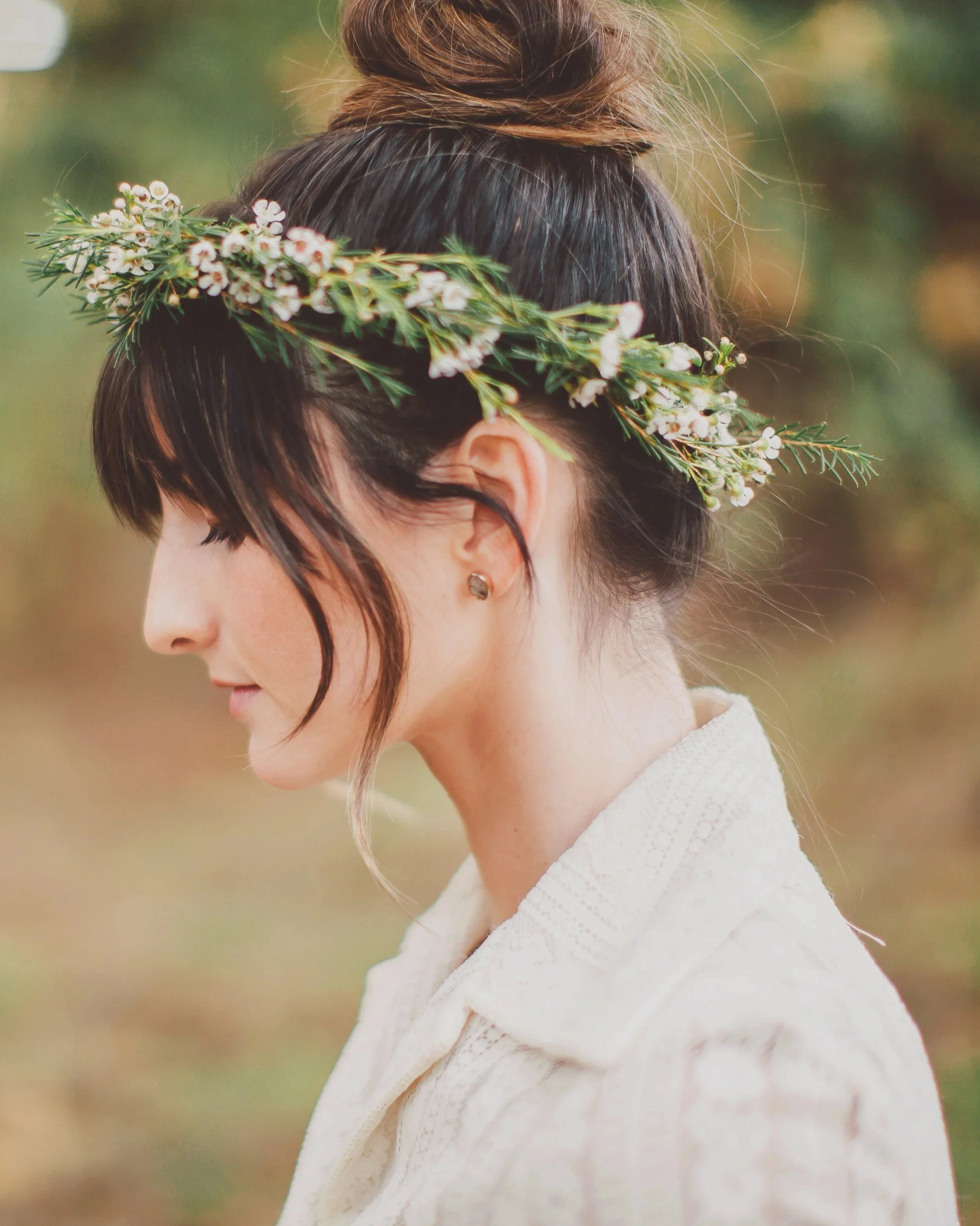 Close-up of a woman with dark brown hair wearing a floral crown of small white flowers and green foliage, dressed in a light-colored high-neck dress, in an outdoor setting.
