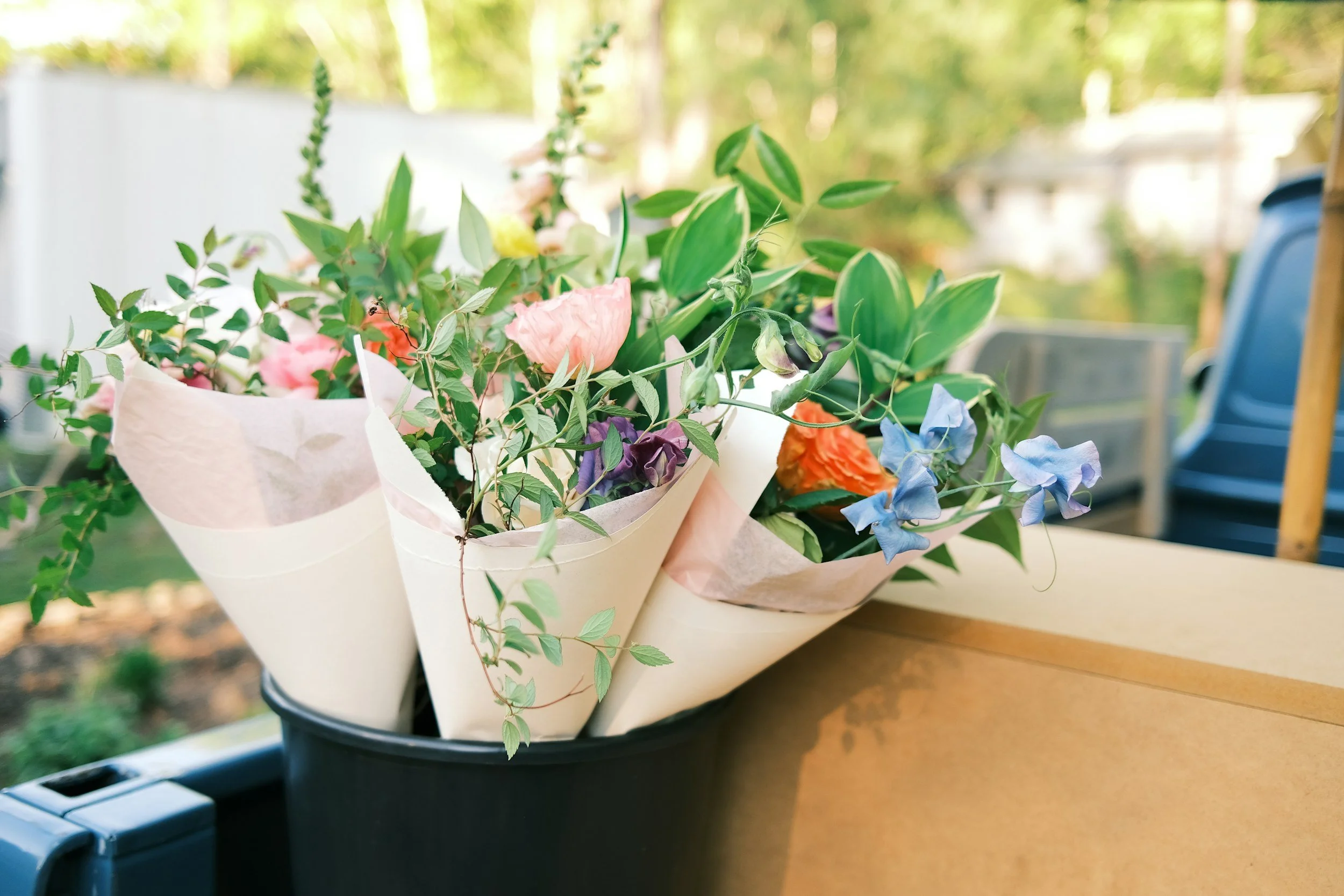 A bouquet of mixed flowers including pink, purple, and orange blossoms wrapped in tissue paper, placed in a black plastic container on a wooden surface, with green foliage and blurred outdoor background.