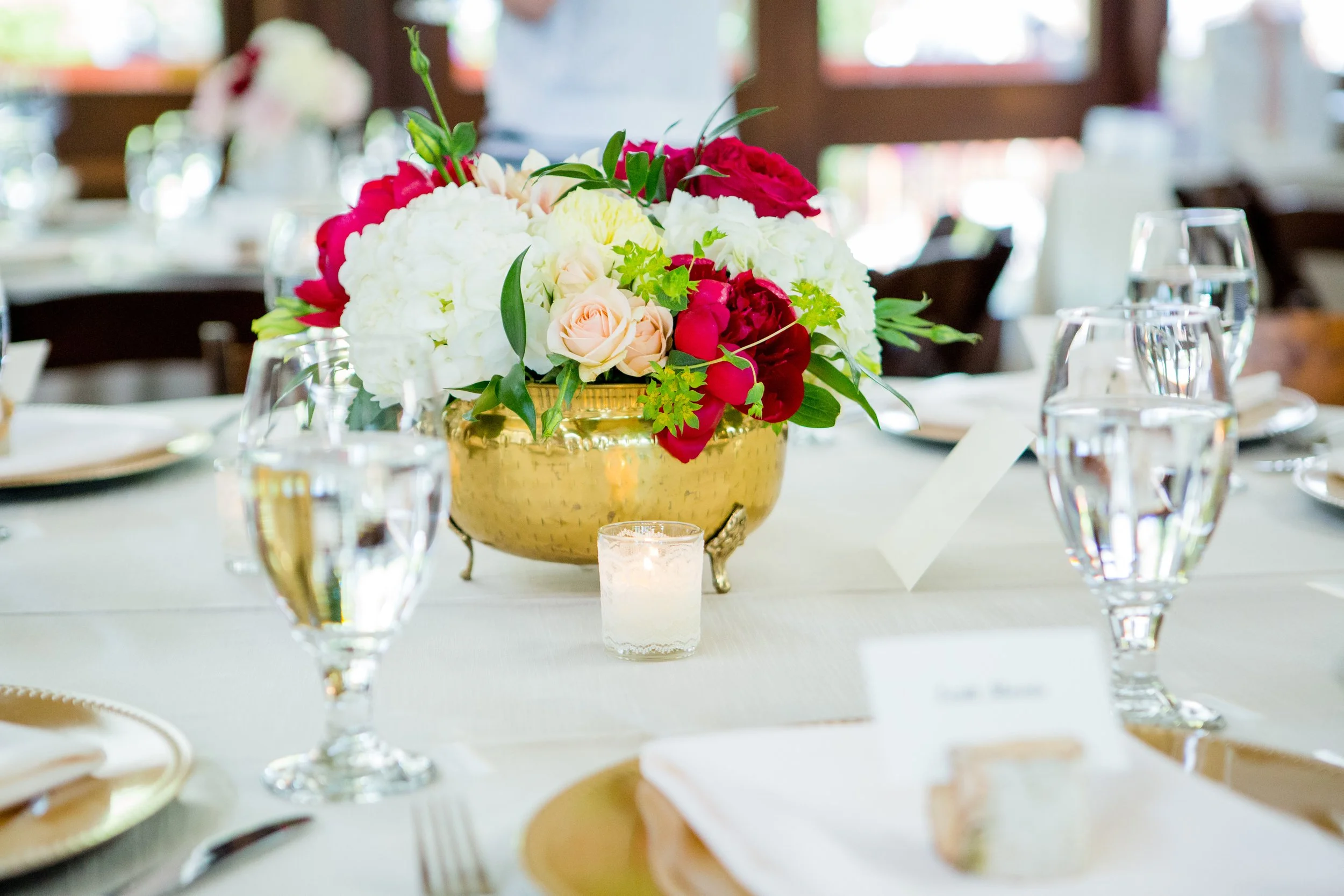 A floral centerpiece with white, red, and blush roses, surrounded by greenery in a gold vase on a table set for an event or celebration.