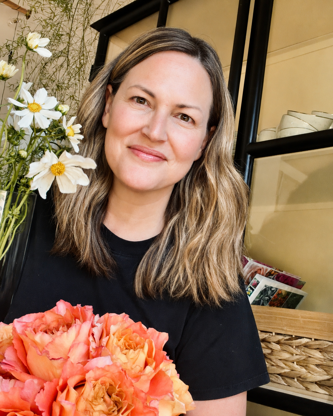 A woman with wavy brown hair holding a bouquet of peach and pink flowers, standing next to a vase of white and yellow flowers, in a cozy indoor space with shelves and a basket behind her.