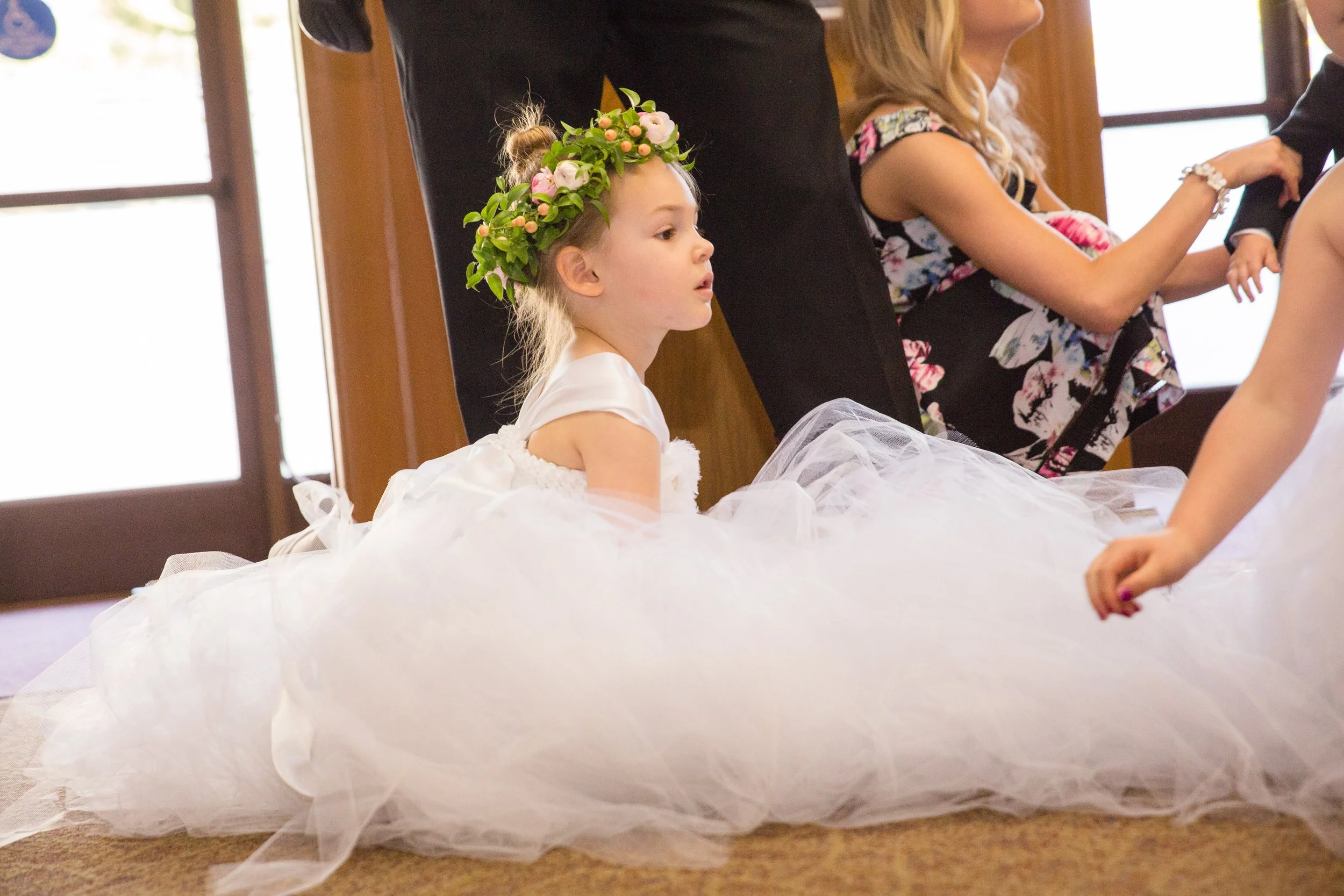 A young girl wearing a floral crown and a white dress sitting on the floor, watching a wedding ceremony.
