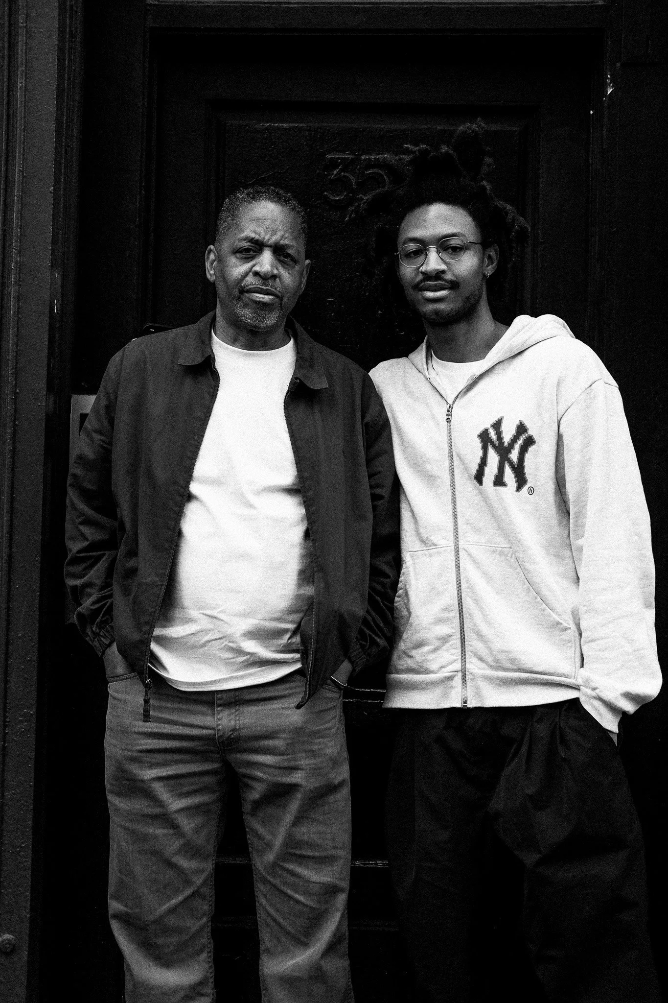 Two men standing side by side in front of a dark door, one older with a serious expression, wearing a black jacket over a white shirt, and one younger with glasses and a hoodie, both with hands in pockets.