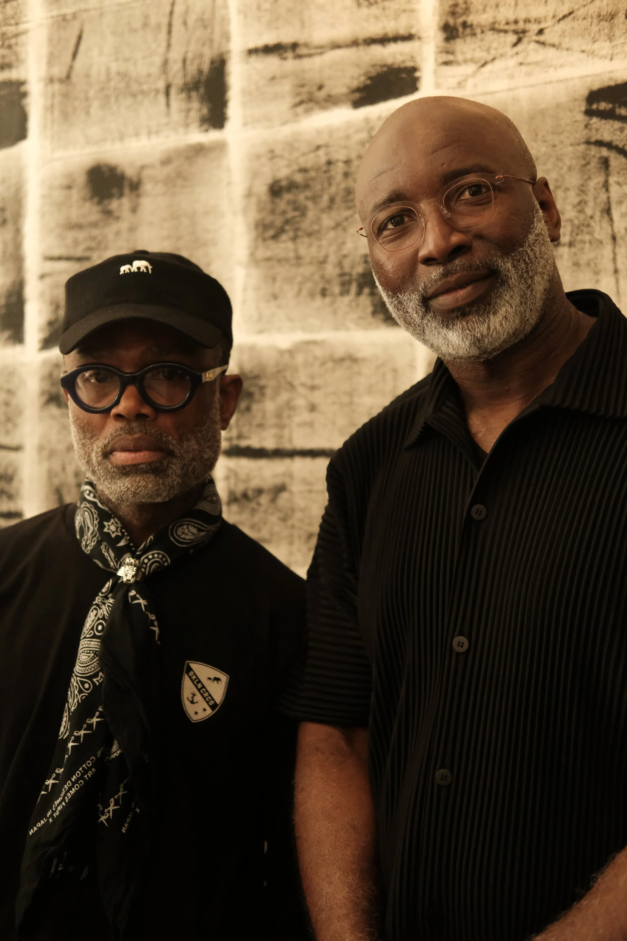 Two men standing in front of a textured wall, looking at the camera. The man on the left wears a black baseball cap, glasses, and a bandana around his neck; the man on the right has a beard, glasses, and is wearing a black shirt.