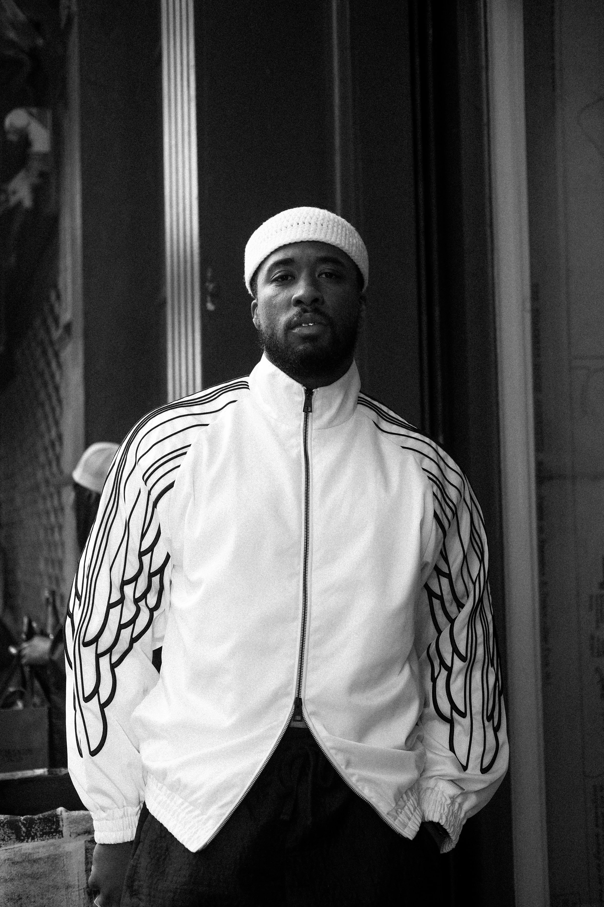 A young man with a beard wearing a white beanie and a white jacket with black line patterns on the sleeves, standing outdoors in front of a building.