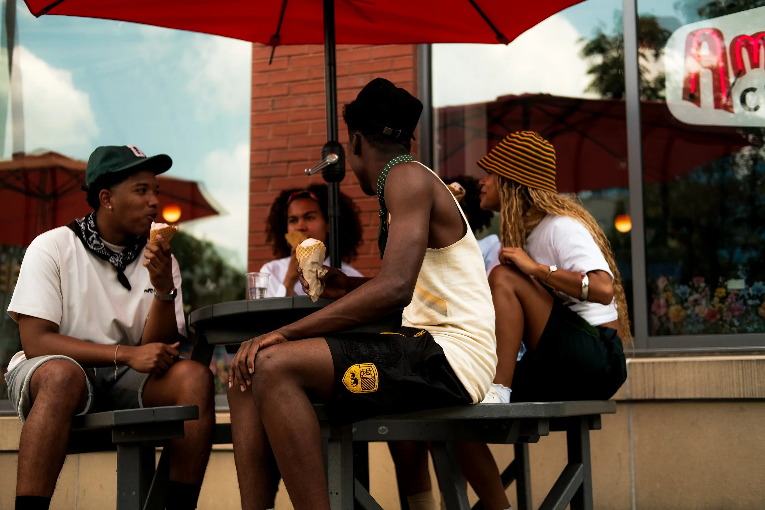 Four young people sitting outside at a table with red umbrellas, eating ice cream and chatting.