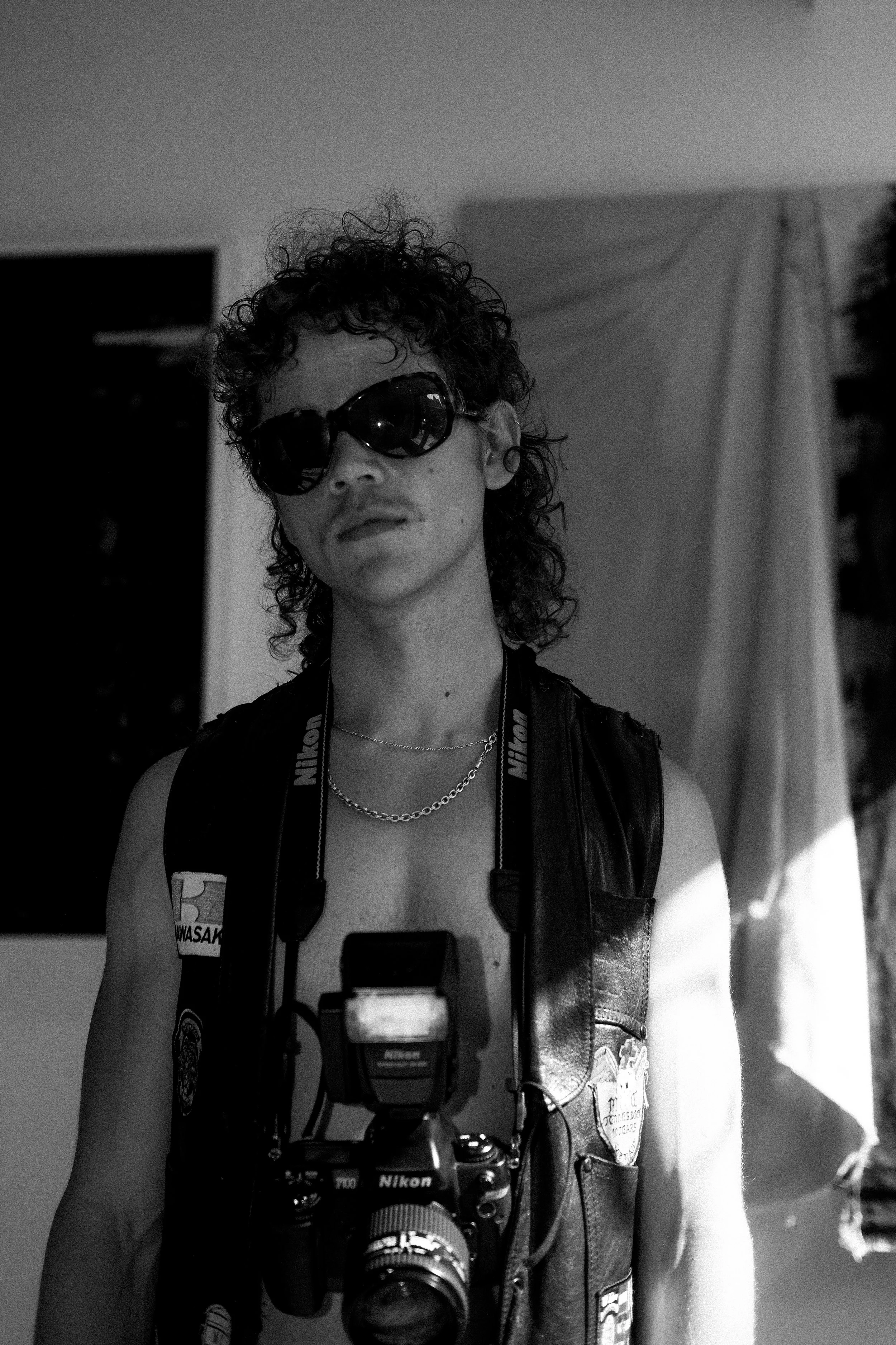A person with curly hair wearing sunglasses, a leather vest with patches, and a Nikon camera around their neck, standing indoors.