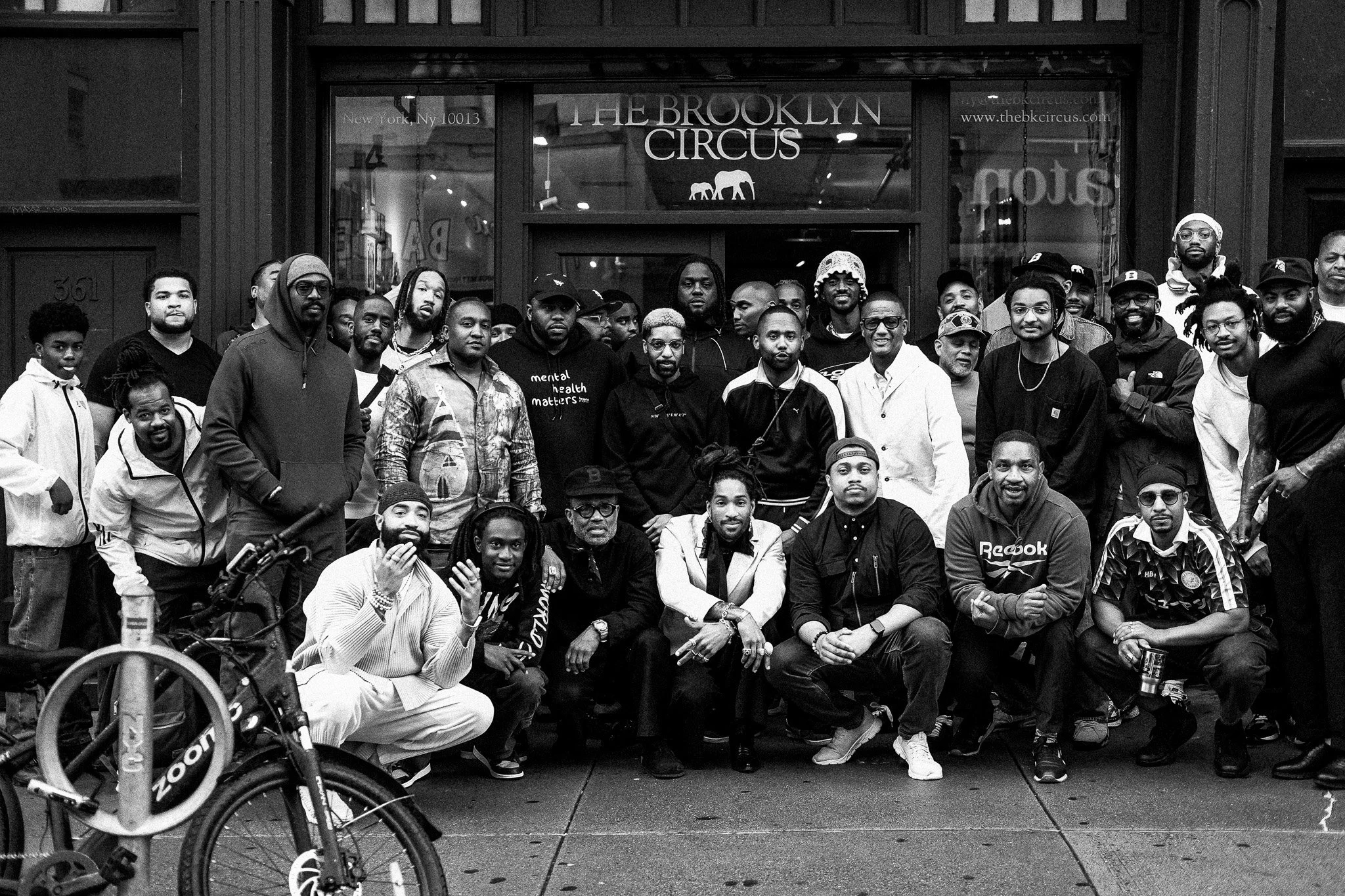 A group of diverse people gathered outside The Brooklyn Circus store on a city sidewalk, black and white photo.