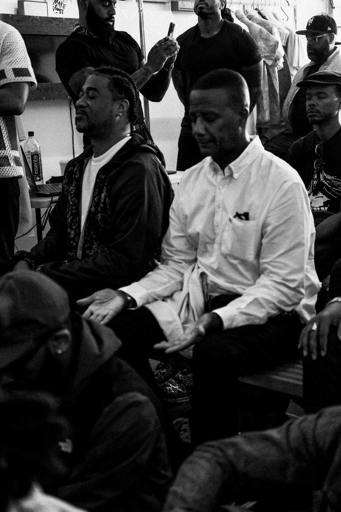 Several people sitting and standing in a room, with two men in the center meditating and others either observing or using their phones.