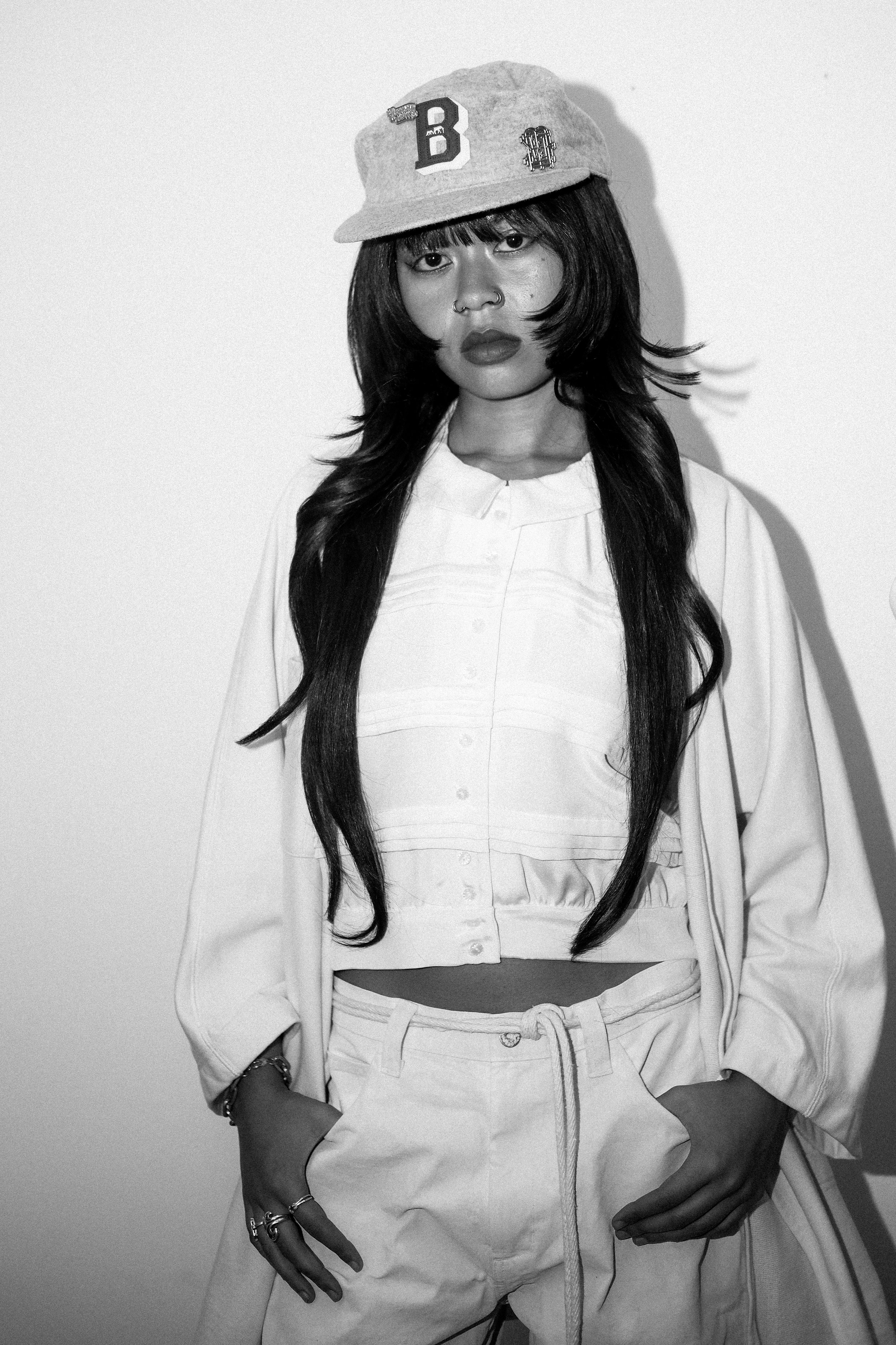 Black and white photo of a young woman with long hair, wearing a baseball cap, a loose jacket over a crop top, and drawstring pants. She has multiple rings and a bracelet, and is posed with her hands in her pockets against a plain background.