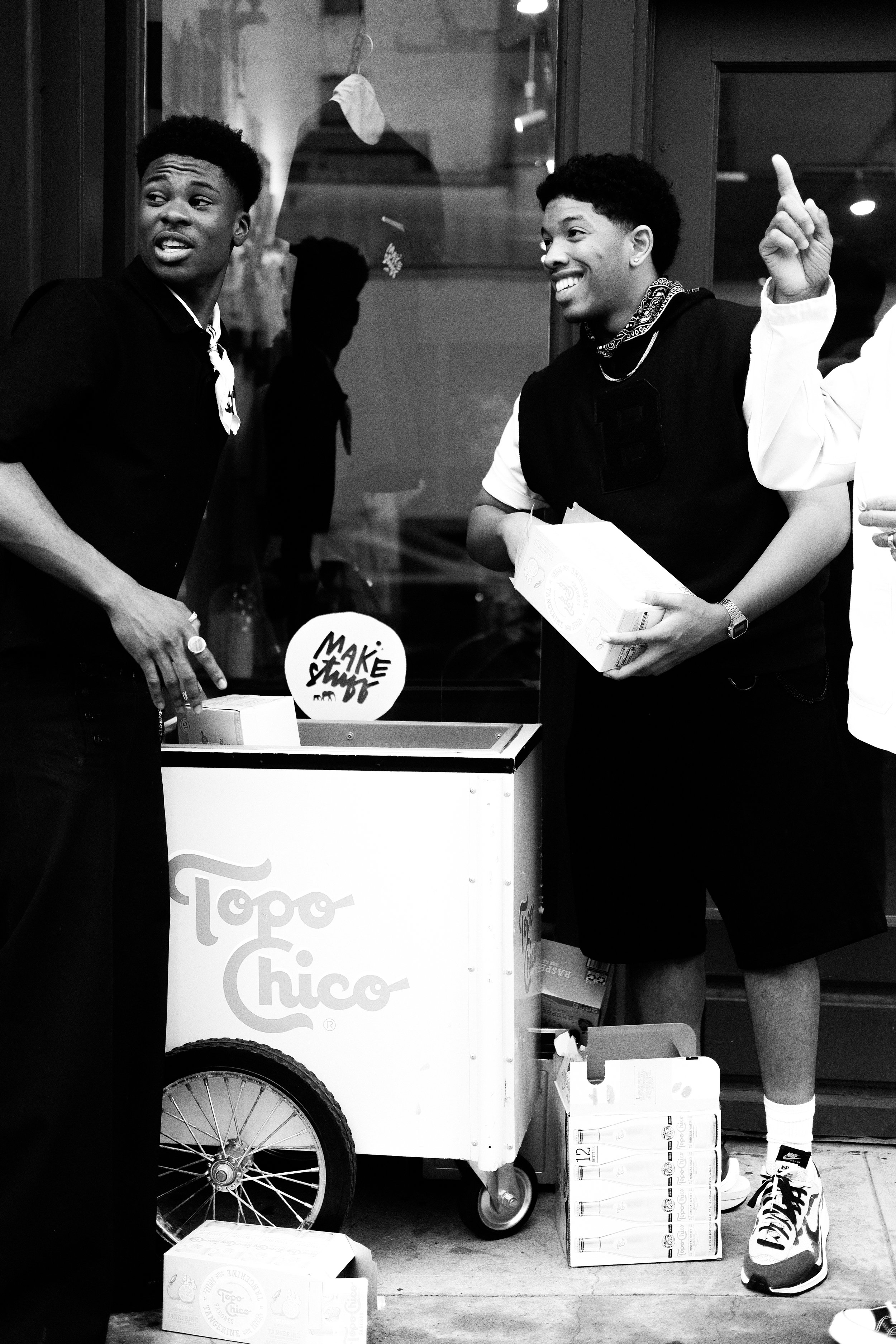 Two young men at a popsicle stand, one holding a box, smiling, and the other looking towards someone off-camera, with a cart that has the sign 'Toog Choco' and a sticker that says 'Make Stuck'.