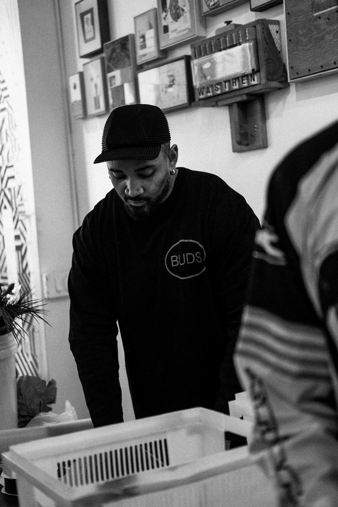 A man wearing a black cap and a black jacket with a "BUDS" logo, leaning over a table in an indoor setting with framed pictures on the wall behind him.