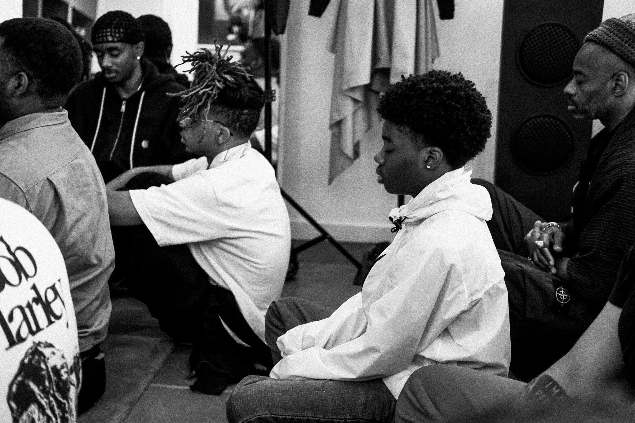A group of people, including young women and girls, sitting on the floor in a room, possibly for a meeting or event, with a speaker in the background.