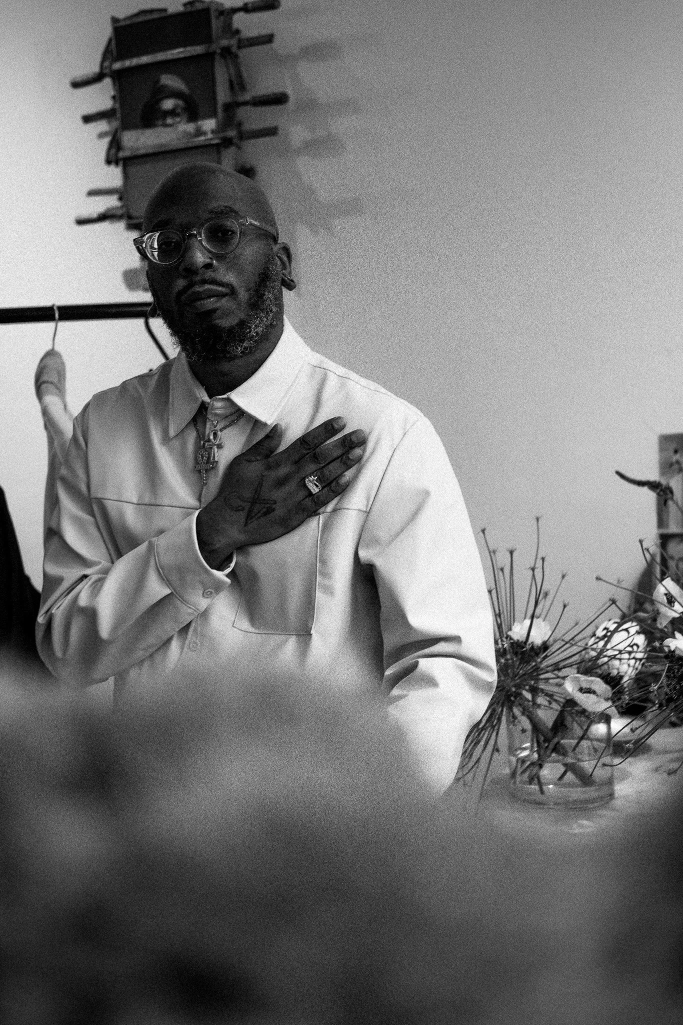 A black-and-white photo of a man with glasses and a beard, wearing a light-colored shirt with a cross necklace, standing indoors with his hand on his chest. There are flowers and some objects in the background.