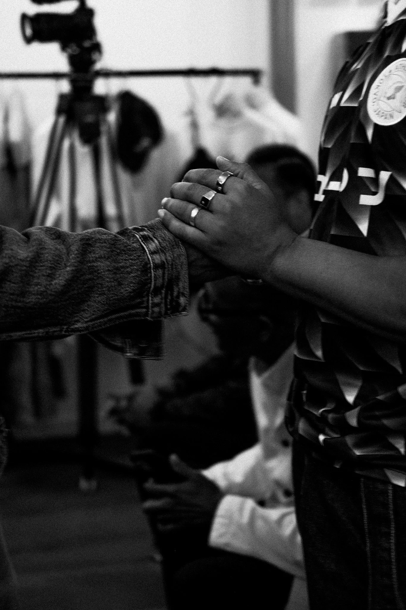 Two people holding hands in a gesture of support or prayer, with a person sitting in the background in what appears to be a church or community setting.