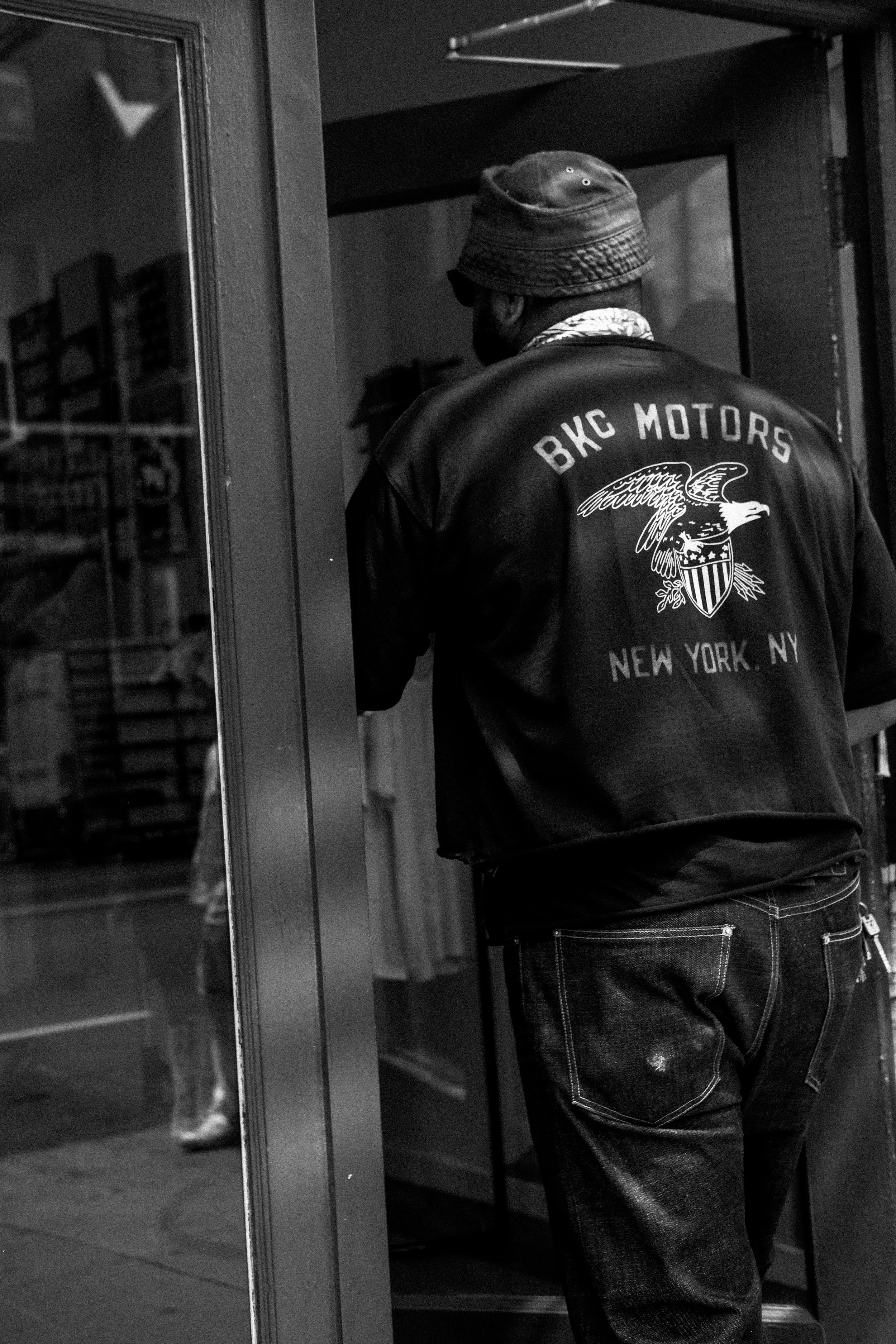 A man in a bucket hat and a leather jacket with 'BKC MOTORS' and 'NEW YORK, NY' printed on the back, standing at the entrance of a shop or building, with his back to the camera.